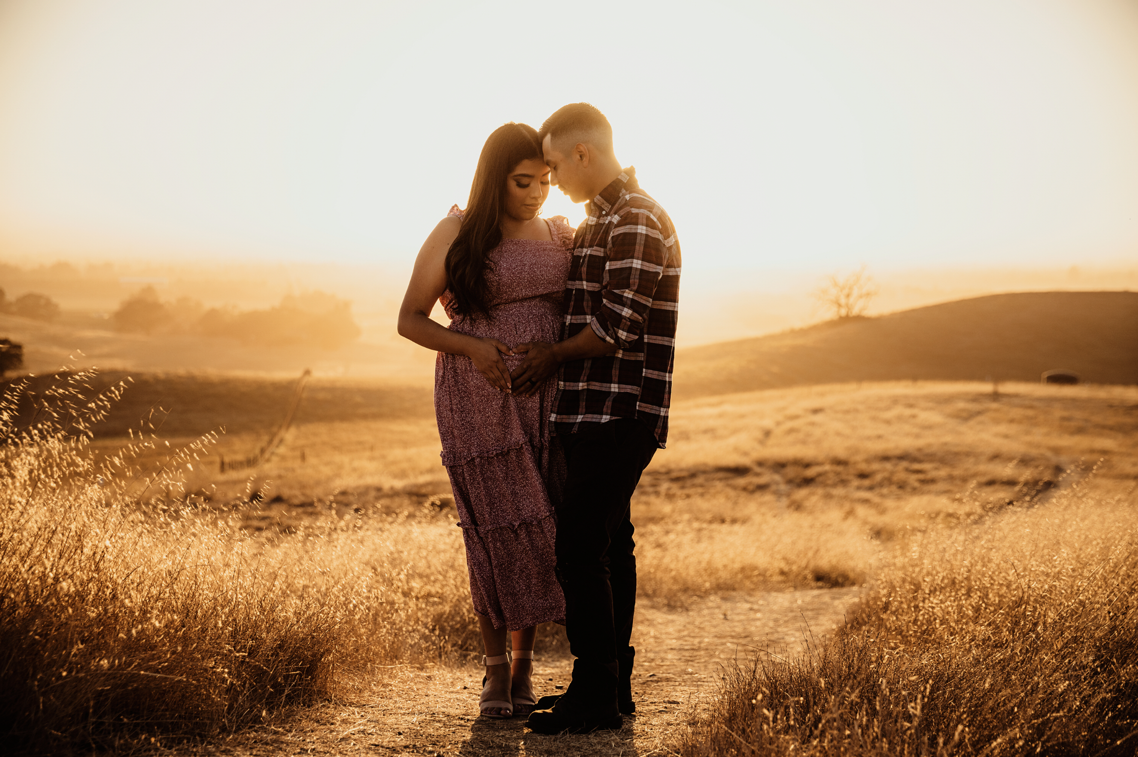 A couple standing in a field during sunset, holding hands and gently touching their foreheads together.
