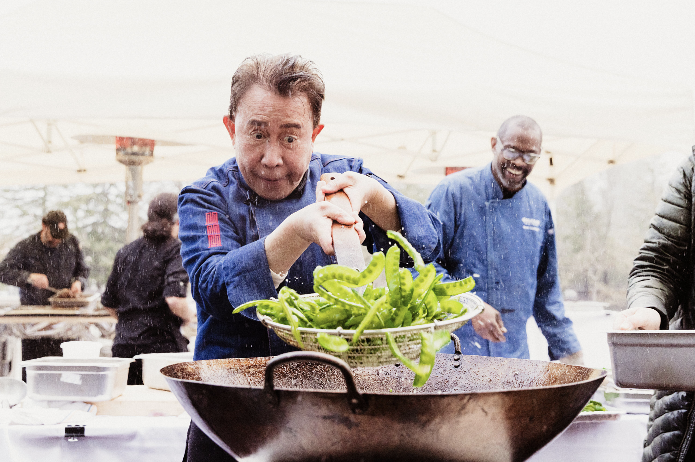 Chef tossing green peppers into a large wok during outdoor cooking event, with other chefs and participants in the background.