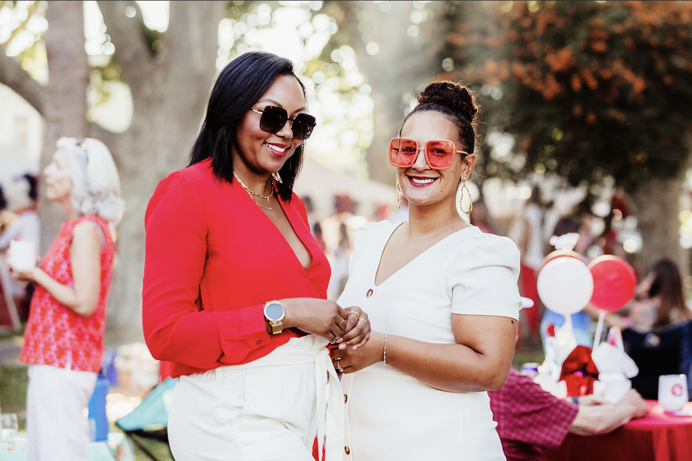 Two women smiling and holding hands at an outdoor event, wearing sunglasses and stylish outfits in red and white, with balloons and people in the background.