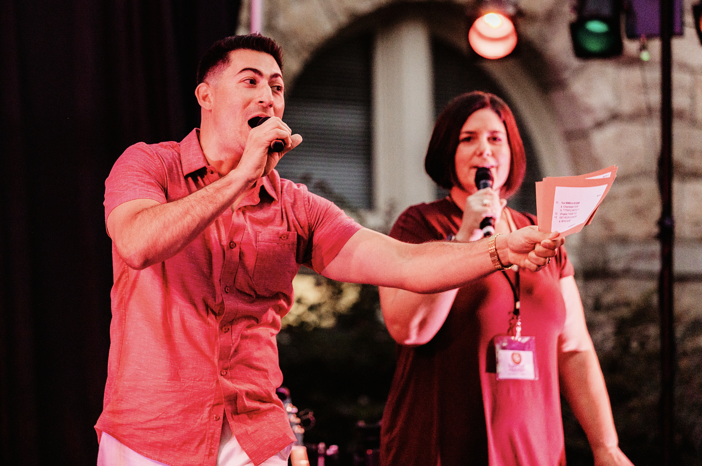 A man and a woman are on stage holding microphones. The man is wearing a pink short-sleeve shirt and the woman is wearing a dark red dress with a lanyard. They appear to be hosting or speaking at an event, with a stone wall and stage lighting in the background.