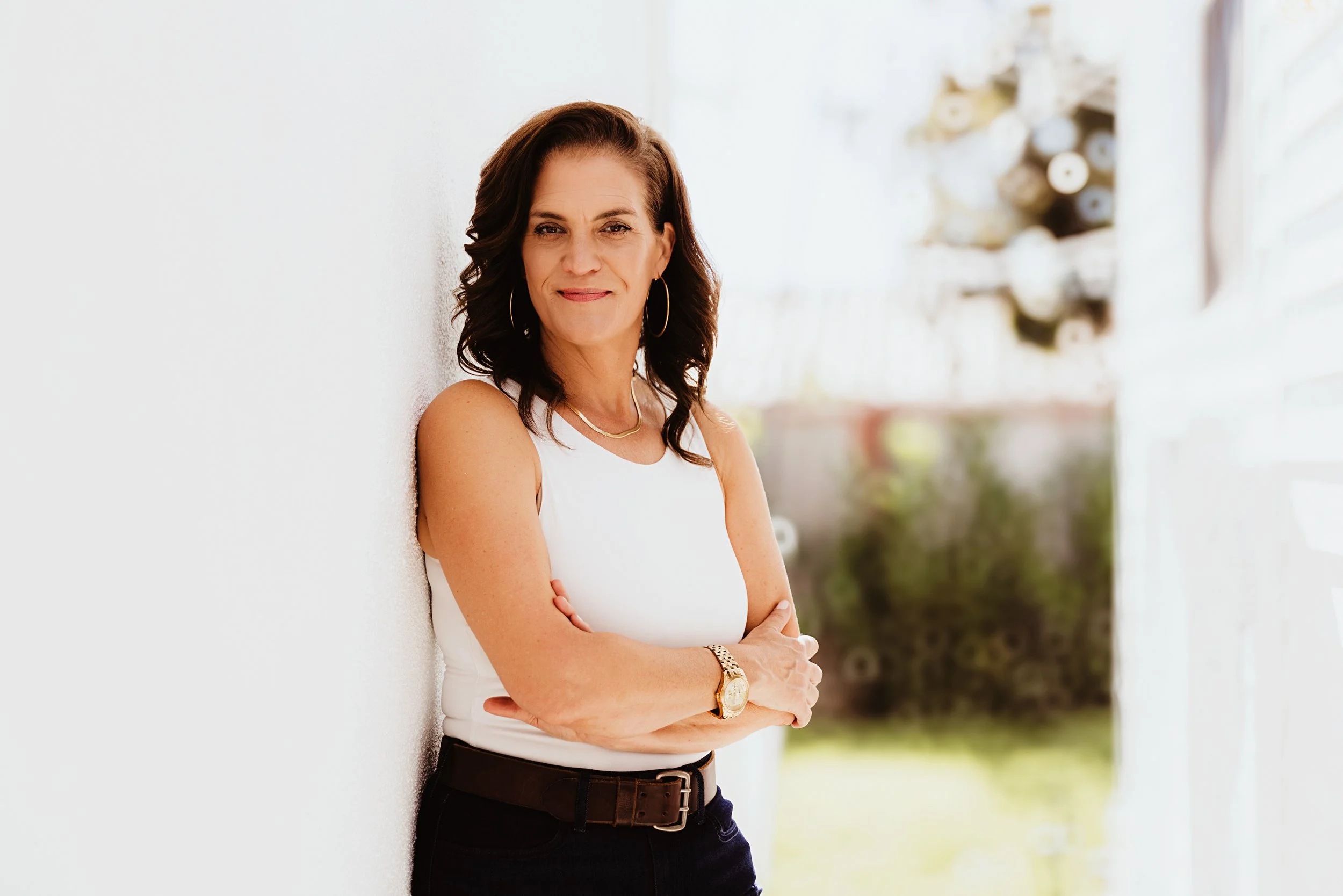 A woman with dark wavy hair, wearing a sleeveless white top, black pants, and jewelry, standing outdoors leaning against a white wall.