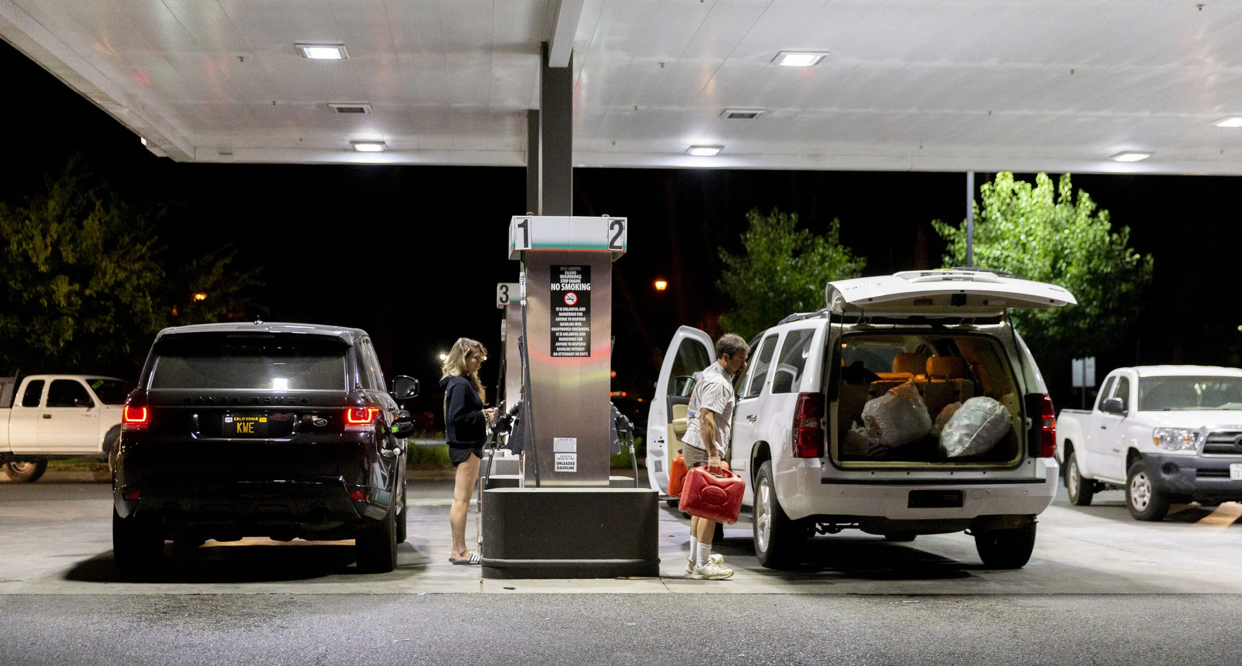 People filling gas at a nighttime gas station with their vehicles parked. One person is a woman with long hair wearing a jacket and shorts, and another is a man with a red gas can. The station has multiple fuel dispensers and a canopy overhead.