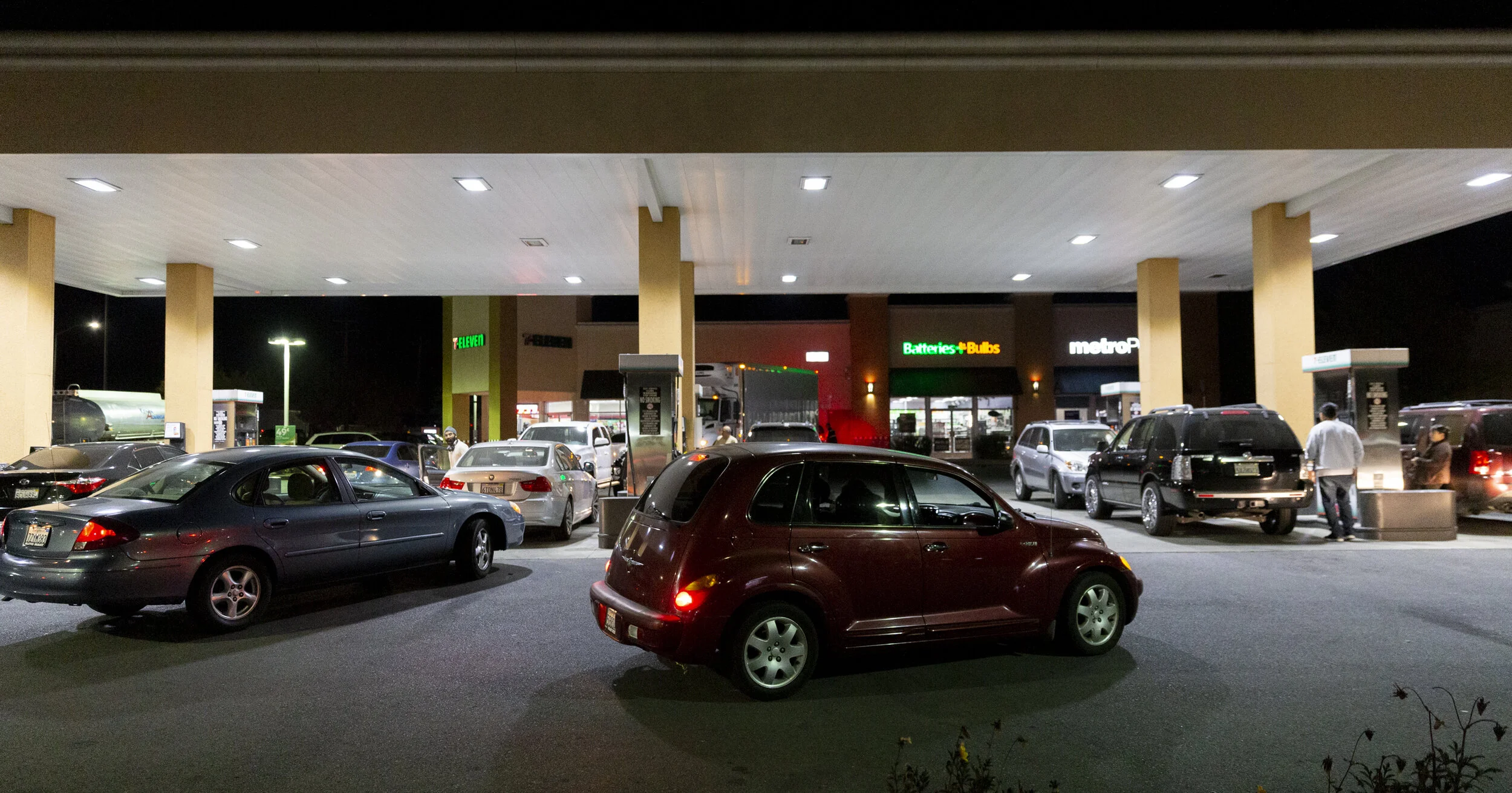 Nighttime scene of a parking lot and gas station with multiple cars parked and people walking. Stores in the background include 7-Eleven, Batteries + Bulbs, and MetroPCS.