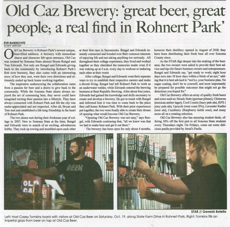 Two men at Old Caz Beer taproom, one pouring beer into a glass, the other standing behind the bar, with shelves of books and beer items behind them.