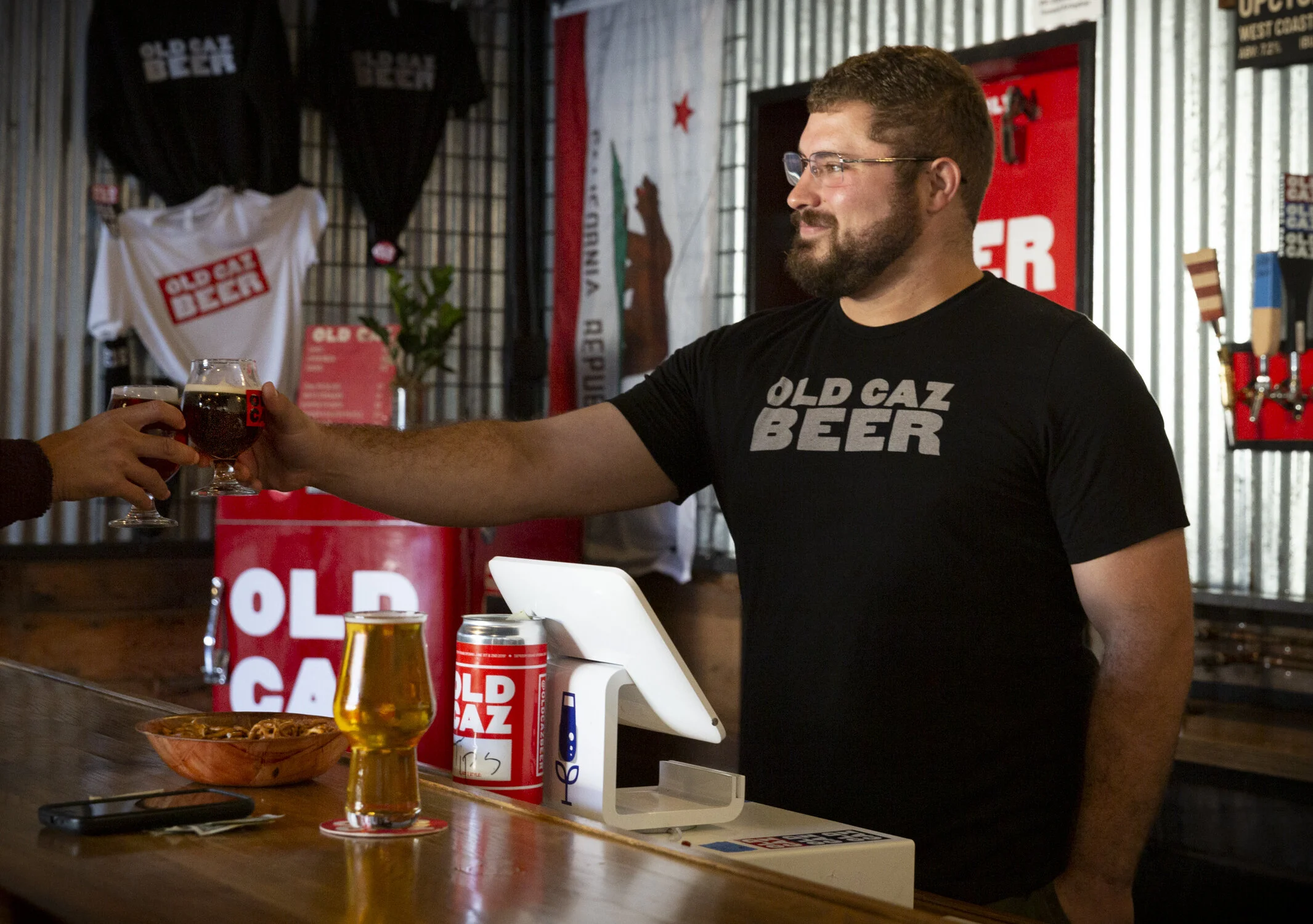 A bartender wearing a black T-shirt with 'Old Gaz Beer' printed on it, serving a dark beer in a small glass to a customer at a bar counter with a cash register, beer cans, and a glass of beer on it. The background has promotional posters and merchand