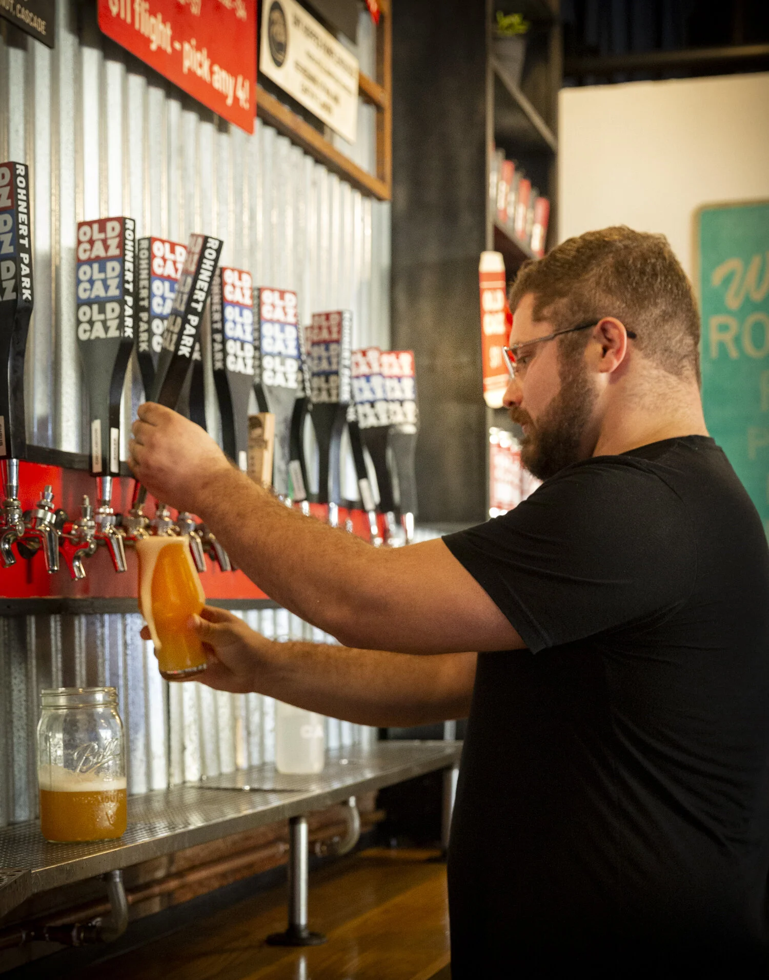 A man with glasses and a beard filling a glass with draft beer from a tap at a bar.
