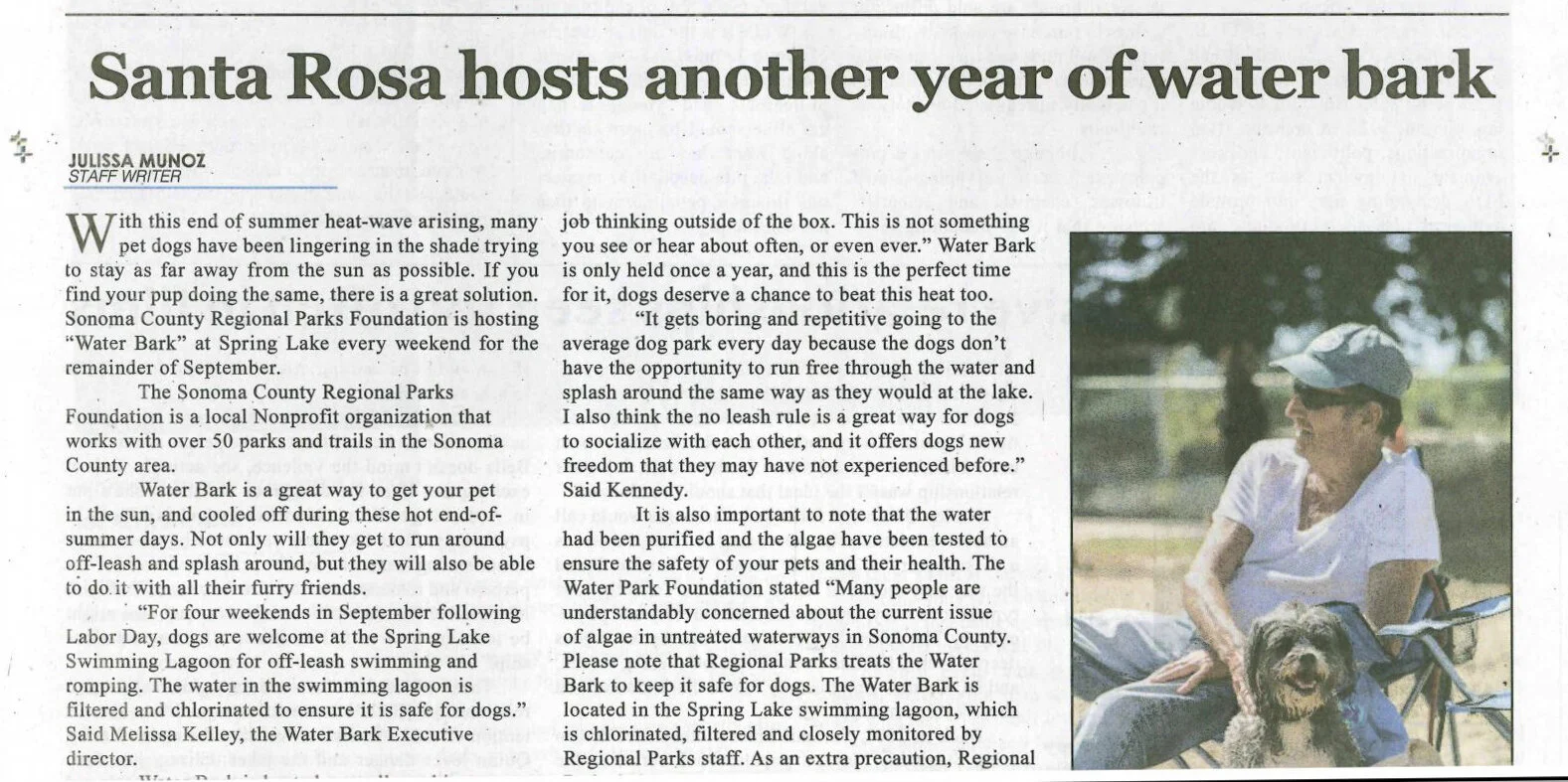 A man sitting outdoors with his dog in a water dog park, wearing sunglasses and a hat, with trees in the background.