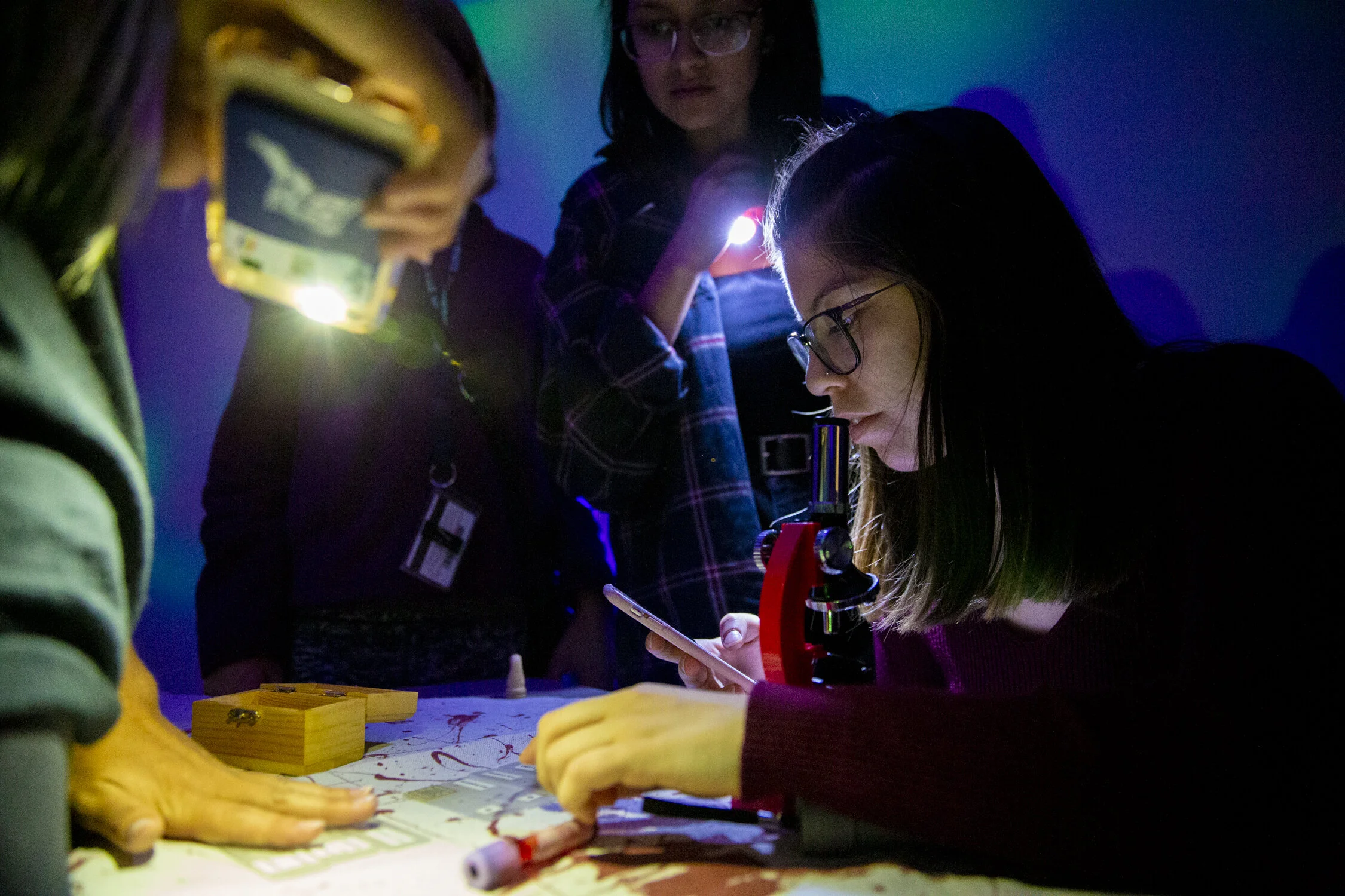Group of young women conducting a science experiment with a microscope, flashlight, and laboratory tools in a dark room with blue and green lighting.