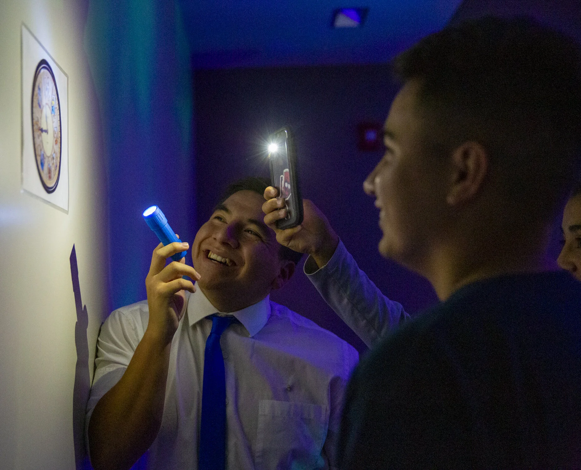 Two young men in formal attire are smiling as one holds a flashlight and the other takes a photo with a smartphone during an interactive exhibit at a museum.