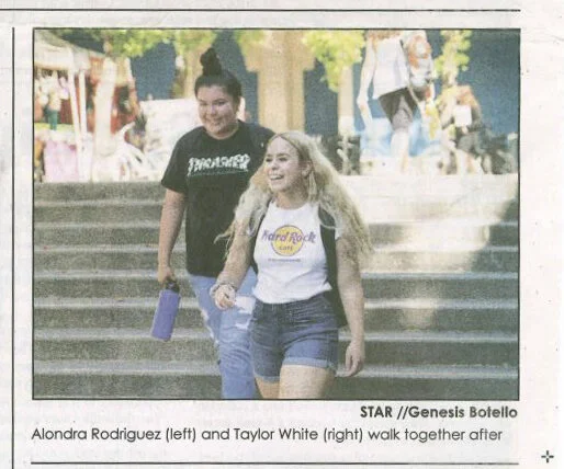 Two young women walking down outdoor steps, smiling and holding hands. One wears a black t-shirt and jeans, the other a white t-shirt with a gold circle logo and denim shorts.