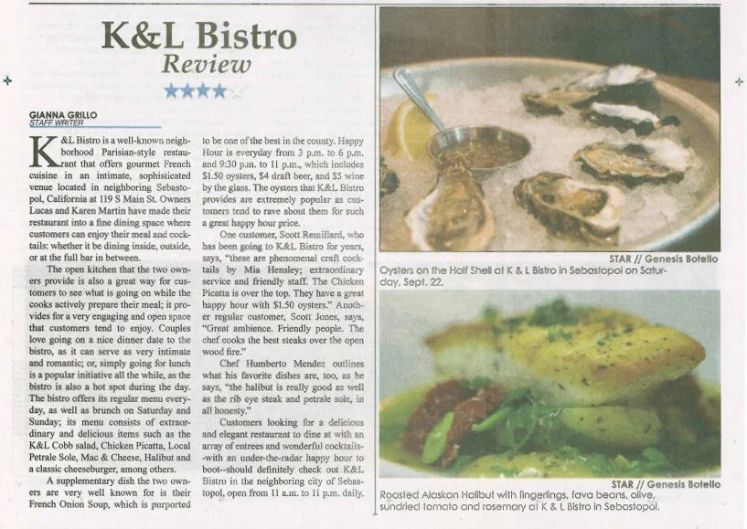 Photograph of a seafood appetizer plate with oysters on the half shell and a roasted salmon dish garnished with lemon, green beans, and tomato, served with sides of olive, sundried tomato, and rosemary, at K & L Bistro in Sebastopol.