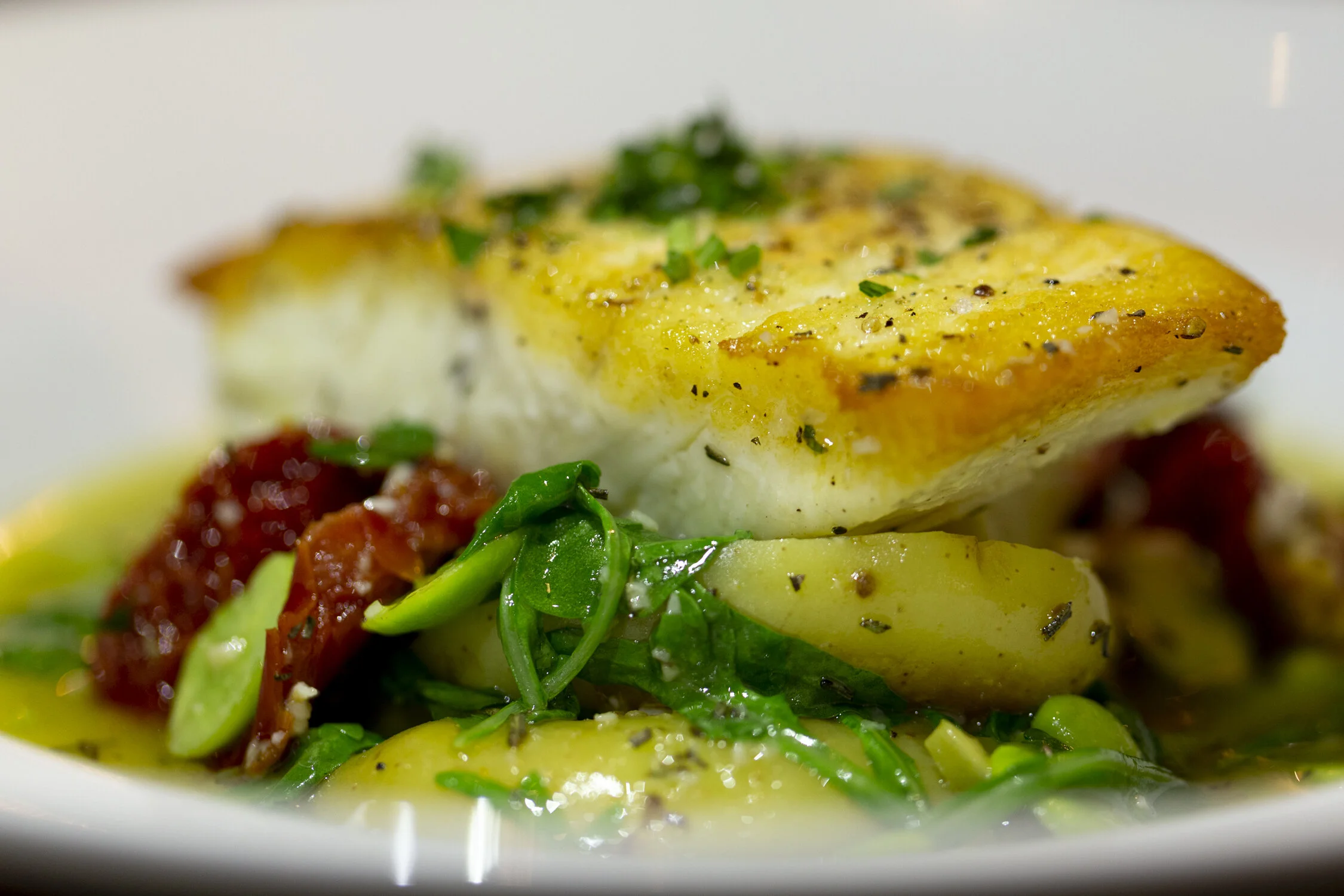 A close-up of a seared fish fillet topped with herbs, served on a bed of green vegetables and potatoes with a tomato-based sauce in a white bowl.