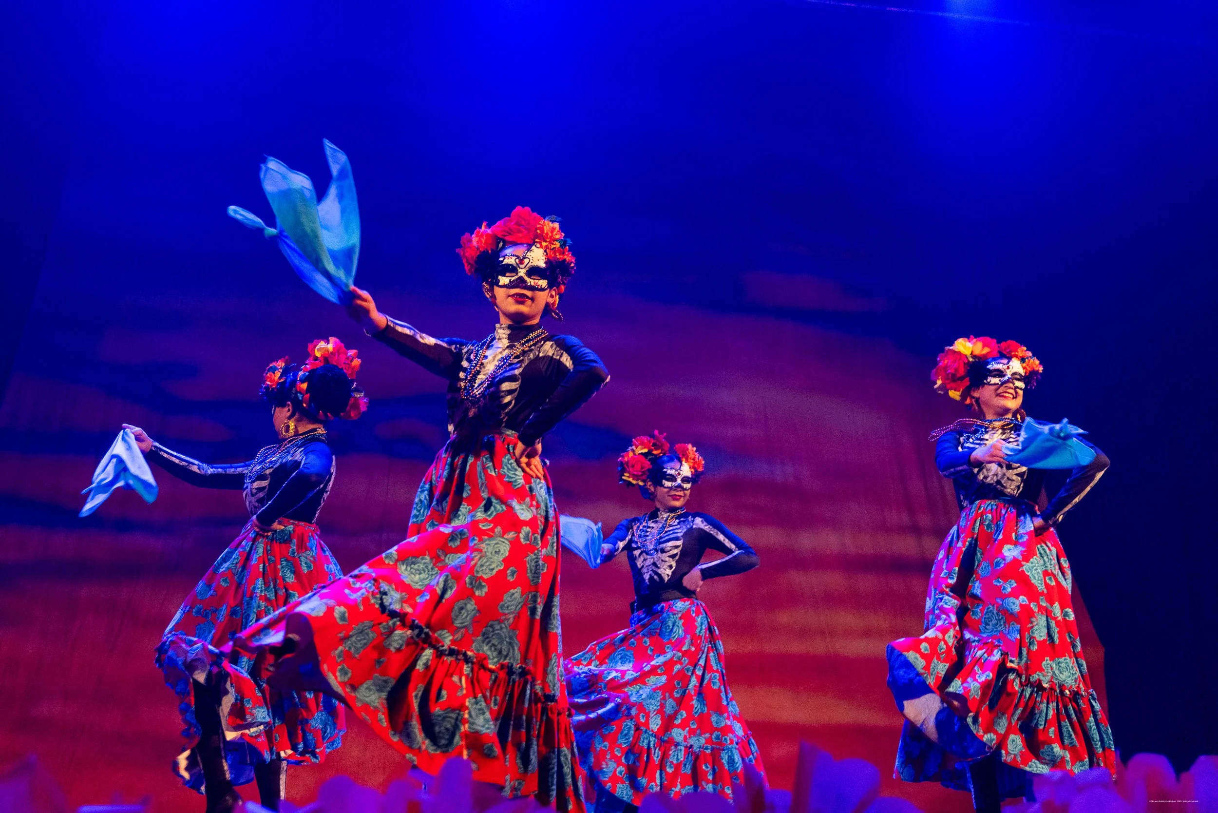 A dancer performing on stage, wearing a colorful costume with large flowing skirts, in front of a red backdrop.