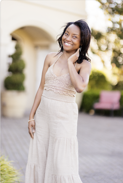 Smiling woman with dark hair and a beige sleeveless dress standing outdoors in front of a building and greenery.