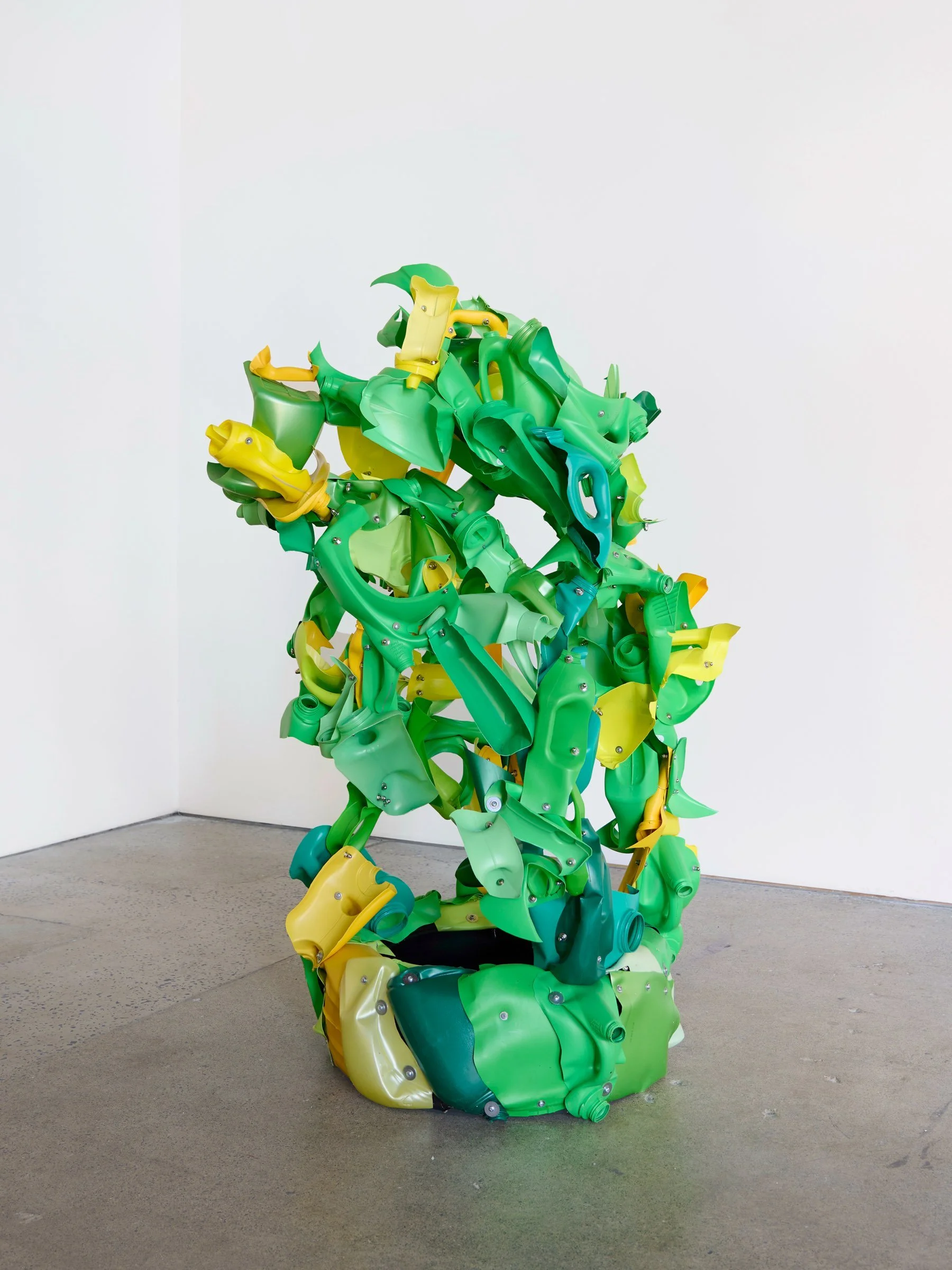 Colorful sculpture made of green, yellow, and blue plastic parts arranged in a humanoid form, displayed in an art gallery with white walls and gray floor.