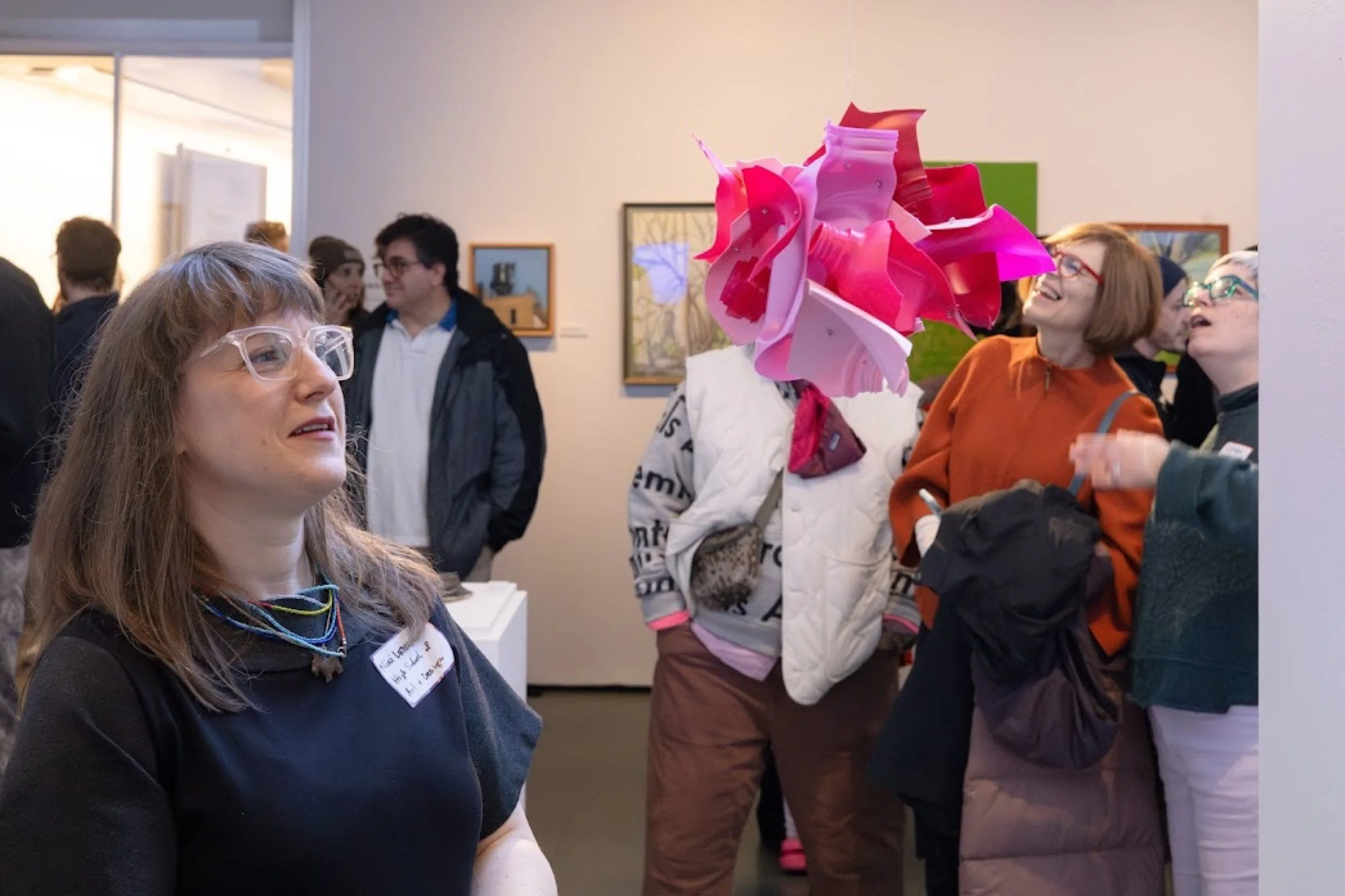 People in an art gallery observing and discussing artwork. In the foreground, a woman with long hair, glasses, and a black shirt with a name tag is looking to her left. Behind her, a woman with a short bob haircut, red glasses, and a rust-colored jacket is smiling, while another woman with short hair, glasses, and a dark sweater is talking to her. A person with a large, colorful headpiece made of pink and red materials is also visible.