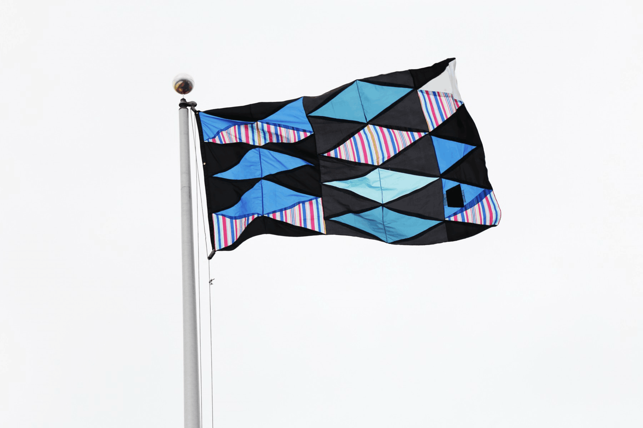 Flag with a geometric pattern of blue, black, and striped sections flying in the wind against a white sky.