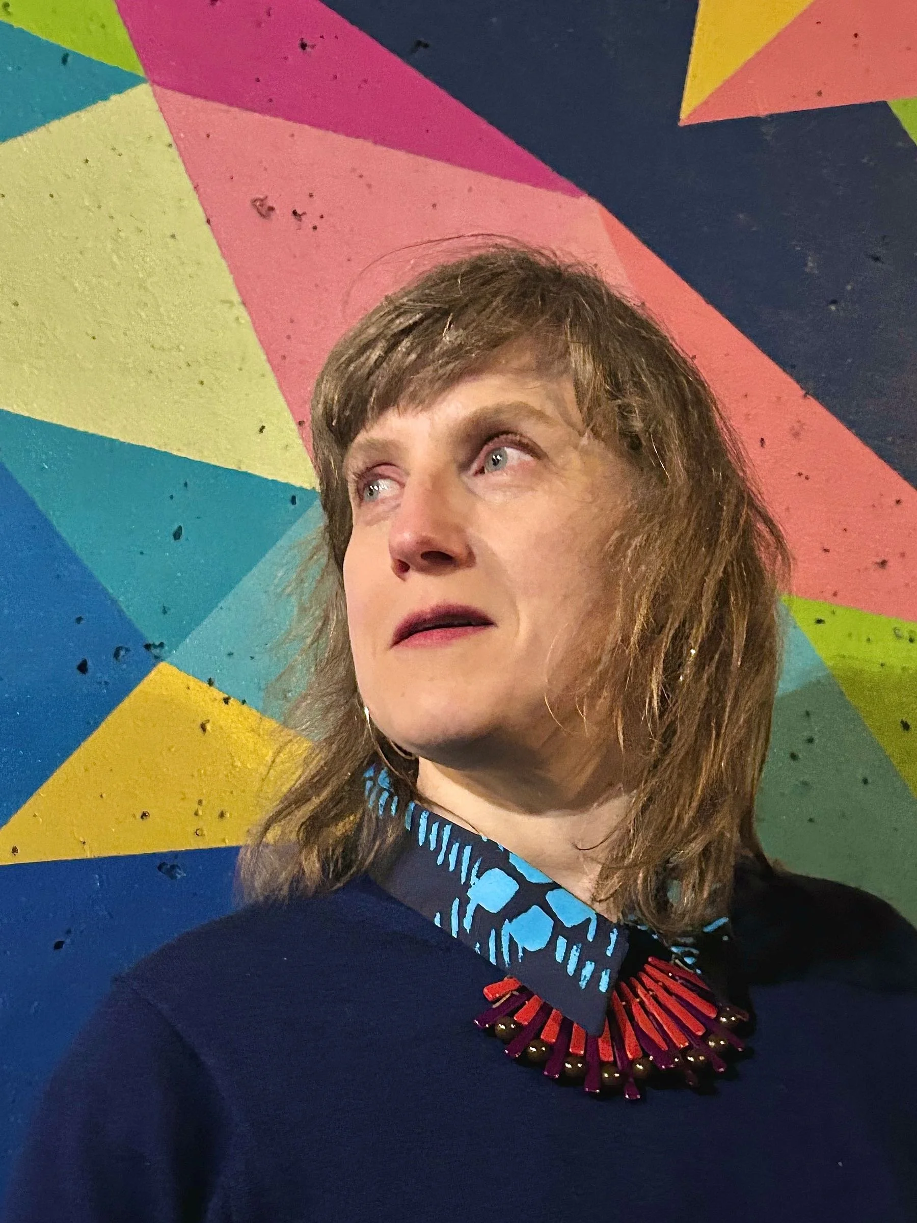 Artist Niki Lederer, wearing a dark blue top with a colorful patterned collar and a statement necklace, standing in front of a brightly colored, geometric mural on the wall.