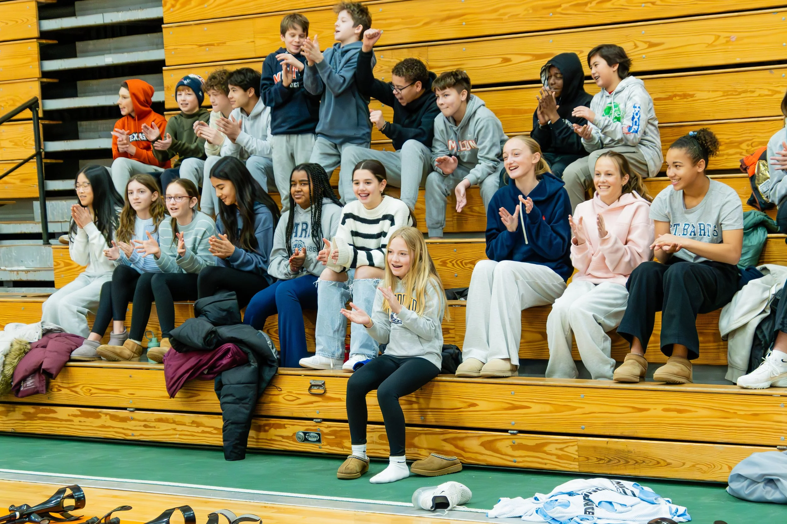 Three people standing on stage, two men and one woman, smiling at an audience of children inside a gymnasium or auditorium. They are wearing casual clothes, with the two men sporting light blue shirts and the woman in a darker shirt. One man has a prosthetic leg and the other is using crutches.