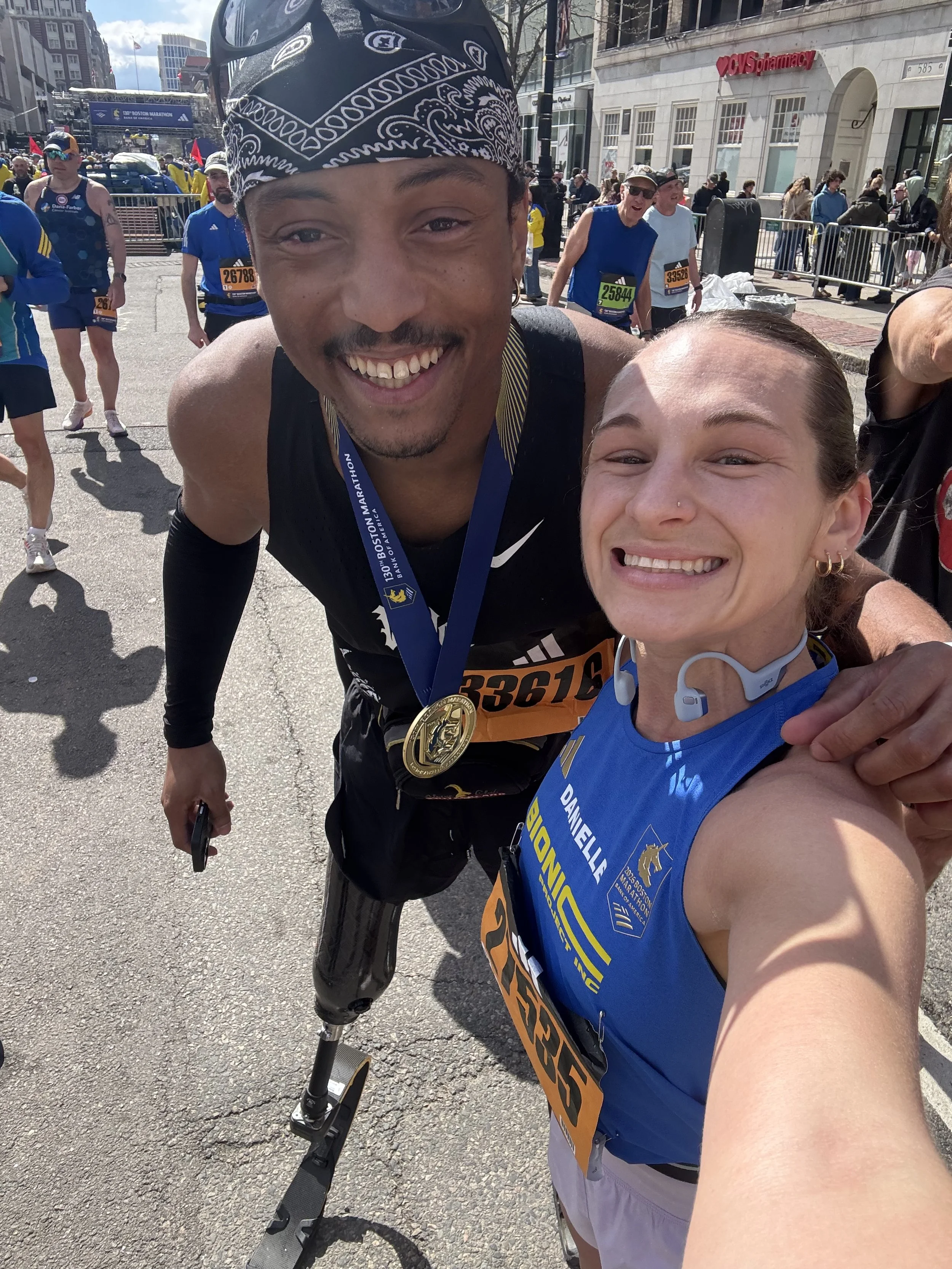 Two smiling Boston Marathon finishers celebrate with their medals, one wearing a Bionic Project jersey and the other using a running prosthesis.