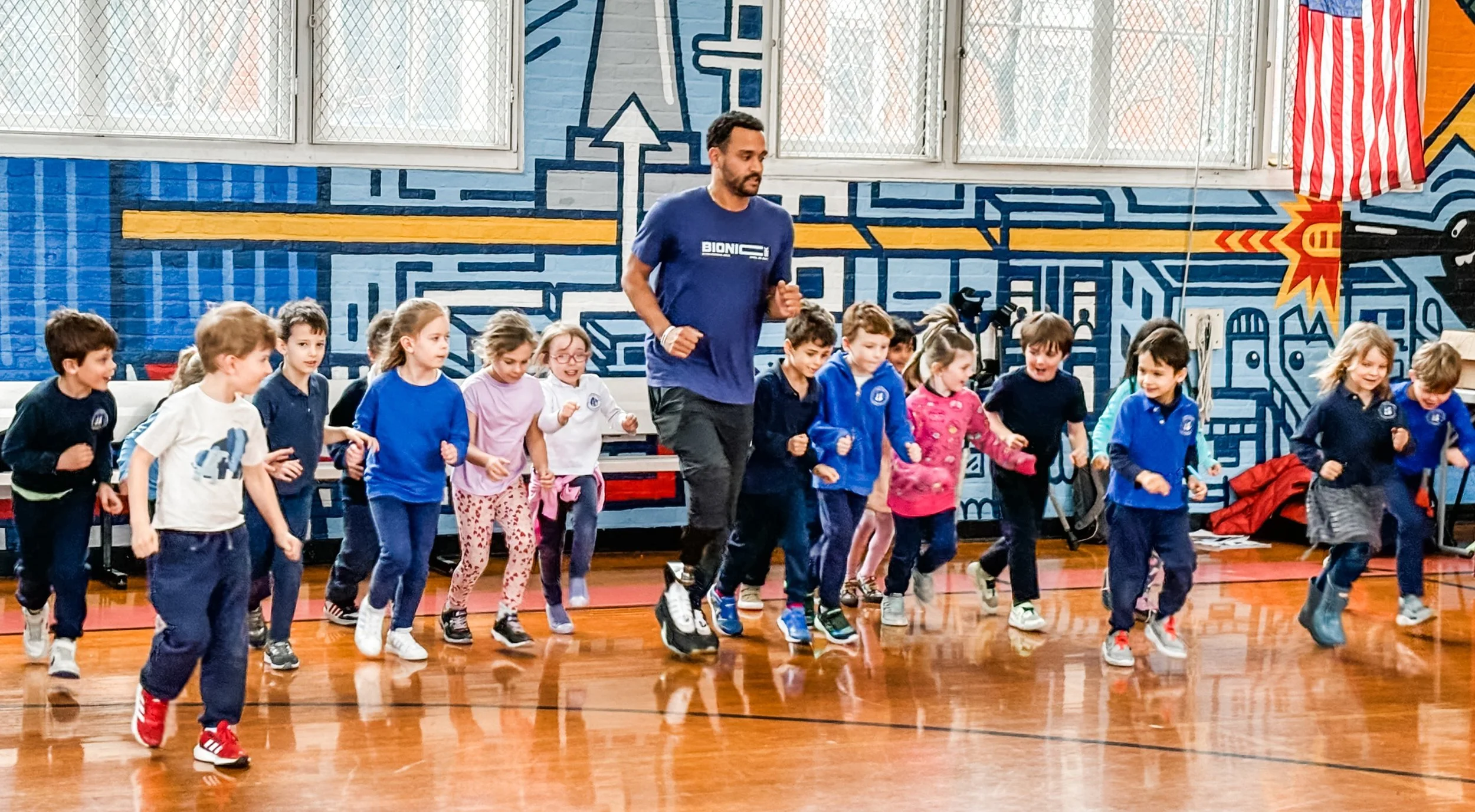 A woman standing in front of a classroom, smiling and pointing to the side. She is wearing a dark gray t-shirt with white text that reads 'BIONICS' and a smartwatch. There are children sitting at desks and colorful posters on the wall behind her.