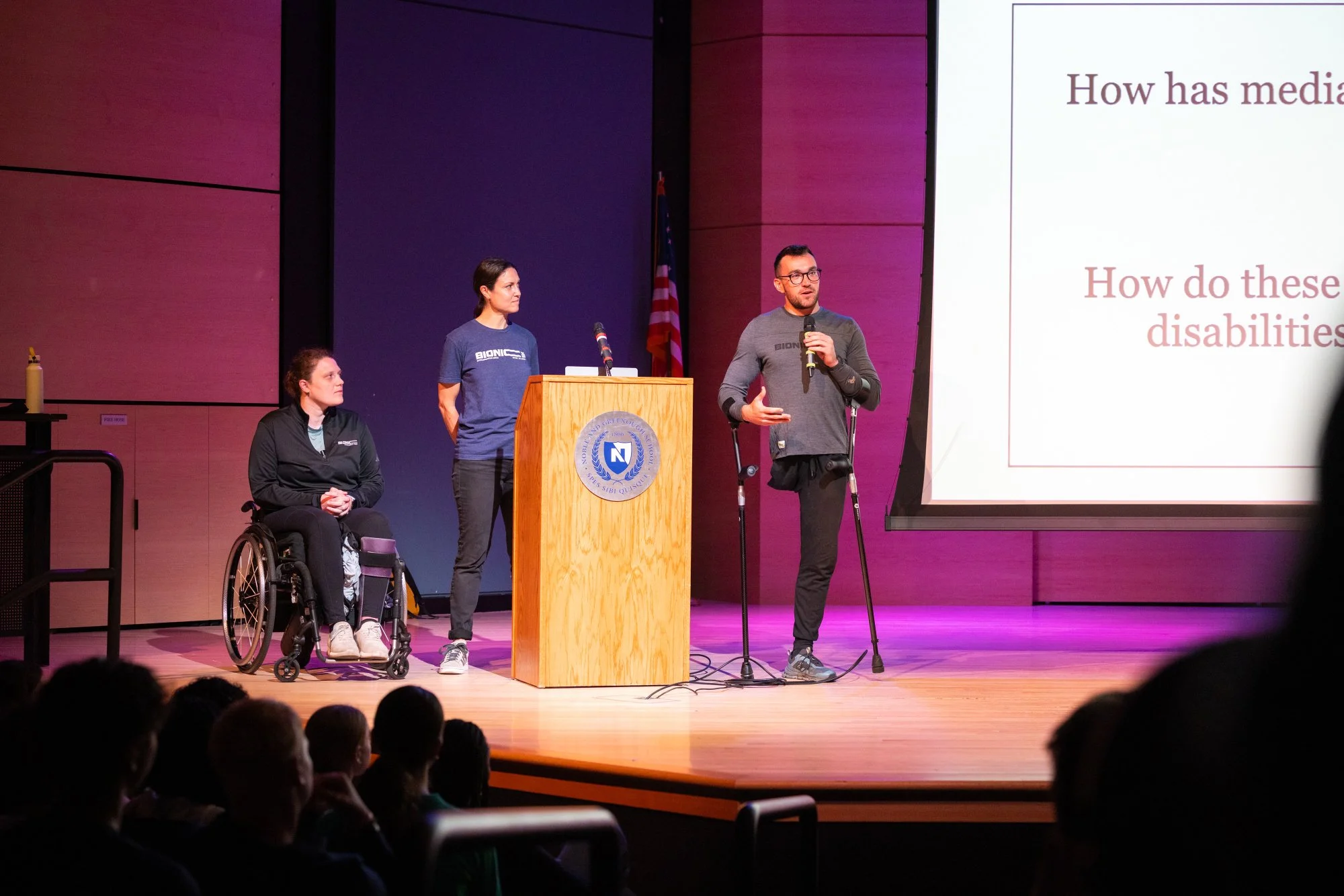 A classroom with students seated around a large table, listening to a male instructor at the front of the room. The classroom has a whiteboard, projector, and educational posters on the walls.