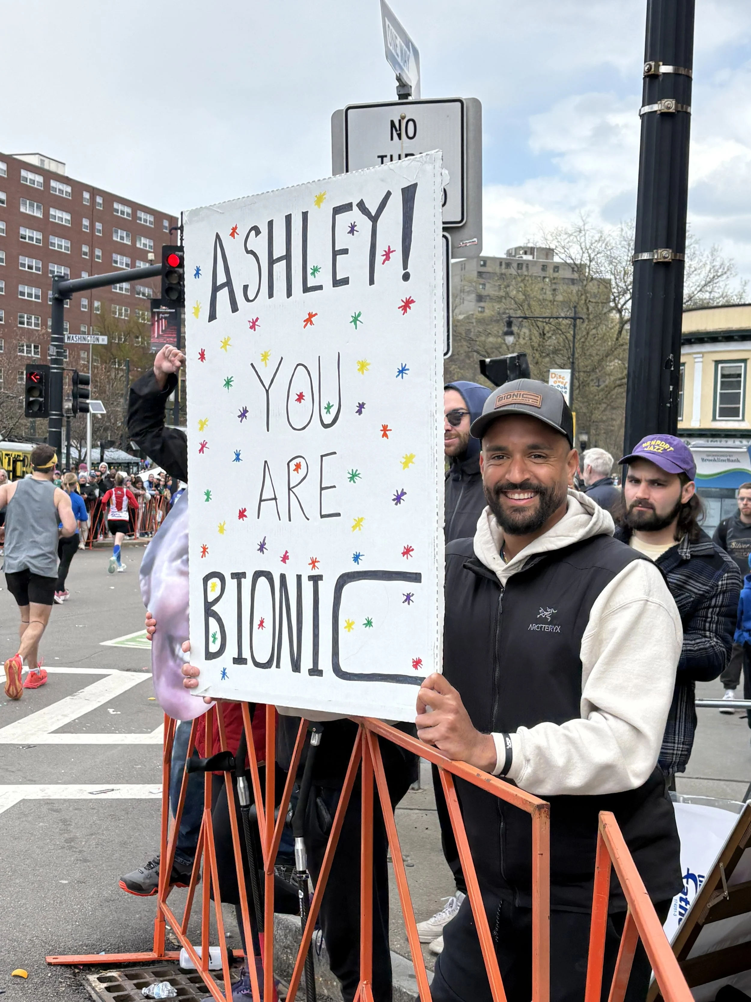A smiling supporter holds a handmade ‘Ashley! You are Bionic’ sign beside the Boston Marathon course.