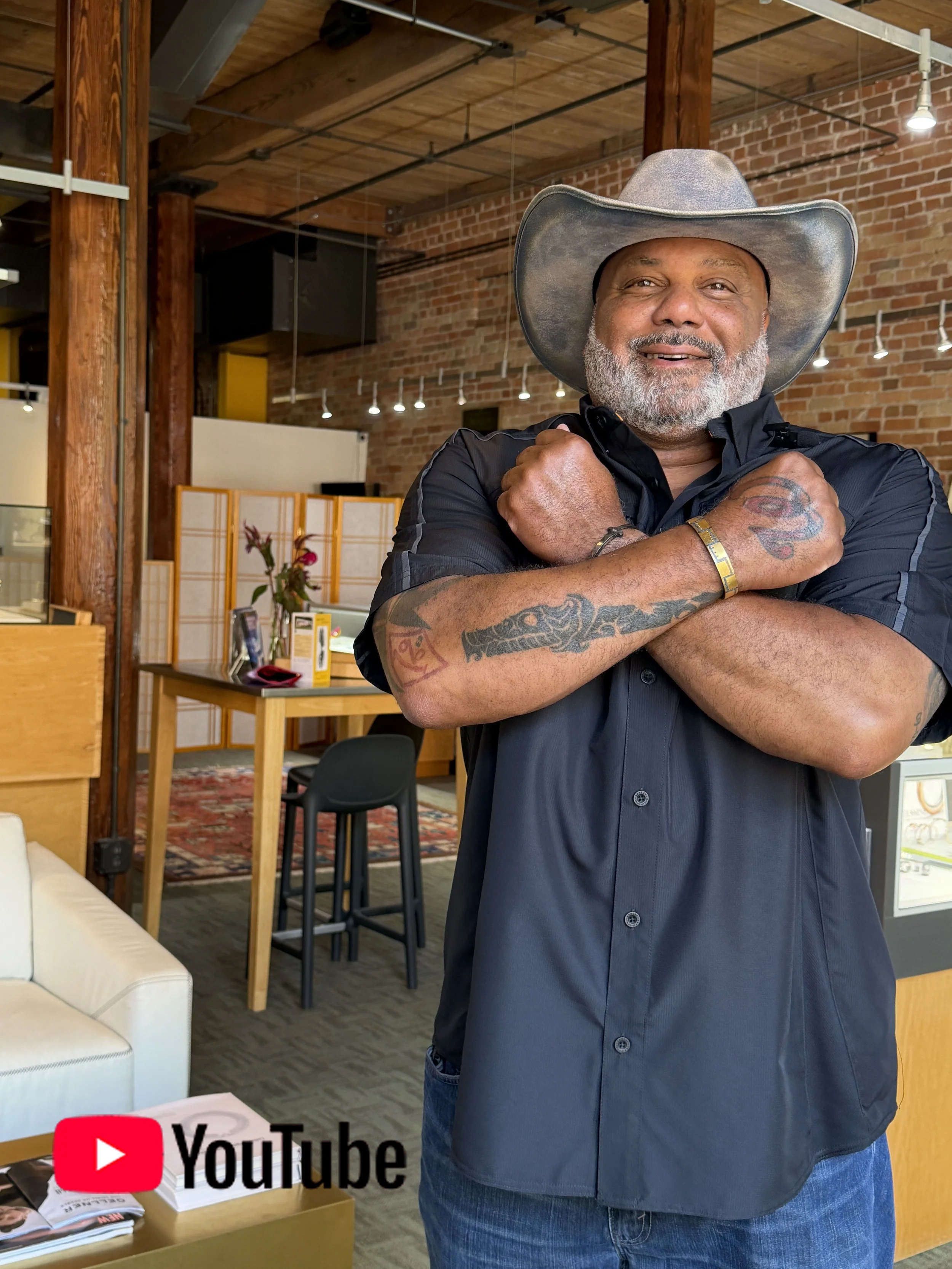 A man with a gray beard and tattoos on his arms, wearing a black shirt and a cowboy hat, crossing his arms and smiling inside a modern brick-walled space with wooden beams and tables.
