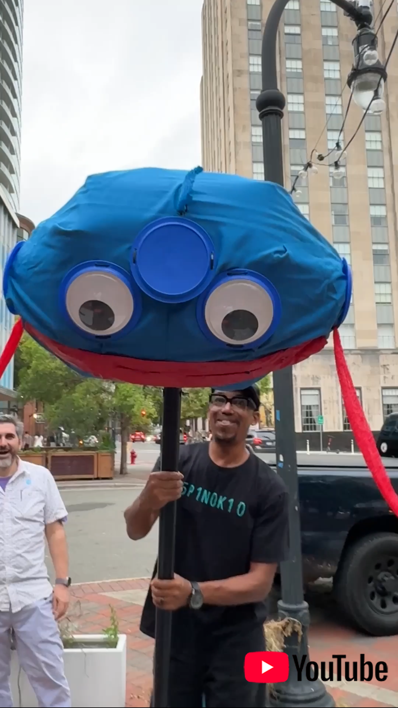 A man smiling and holding a pole with a large blue fish puppet with googly eyes on a city street.