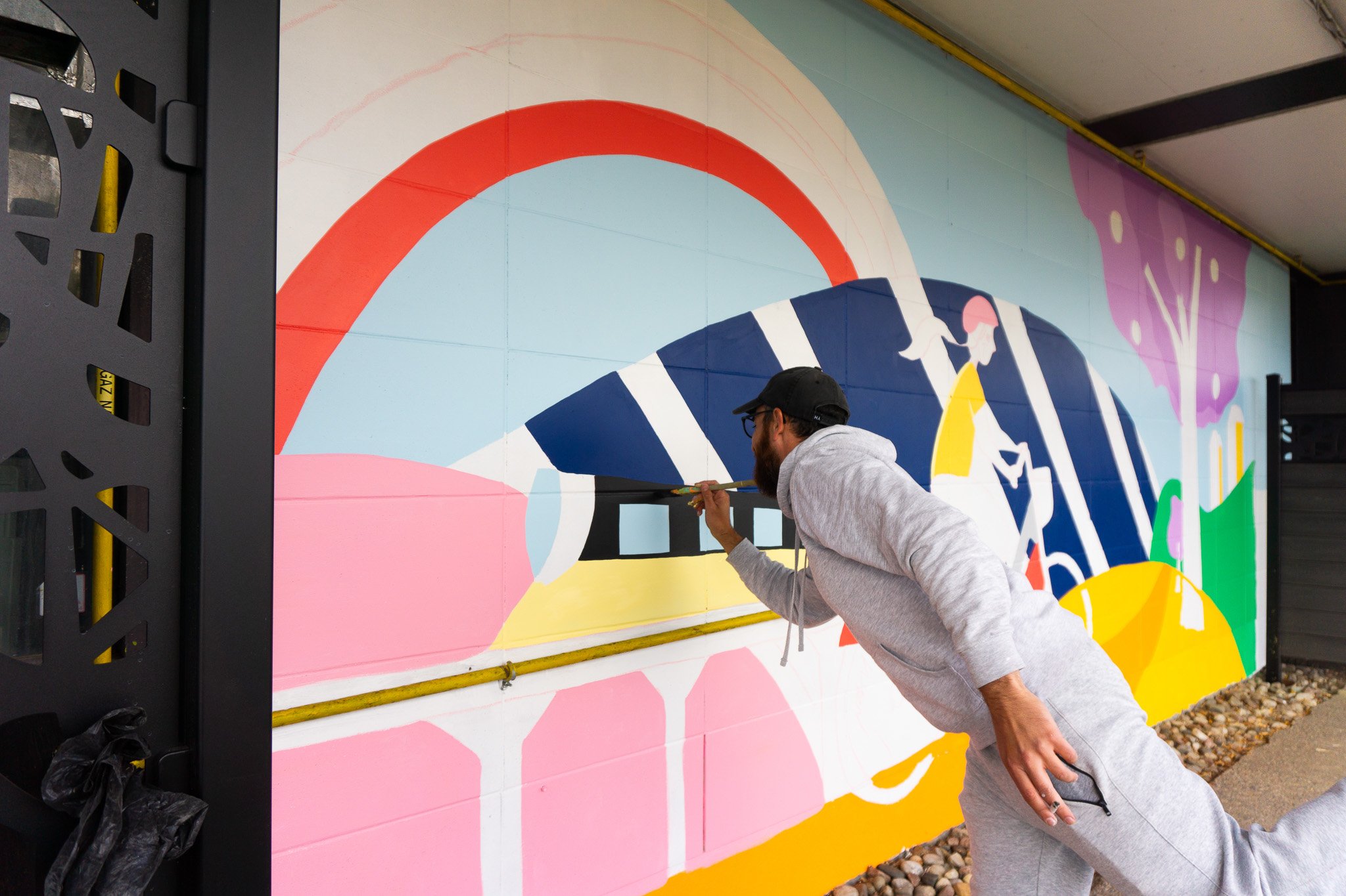 Un artiste peint une murale colorée avec des formes abstraites et des personnages en plein air. Il porte une casquette noire et un sweat à capuche gris.