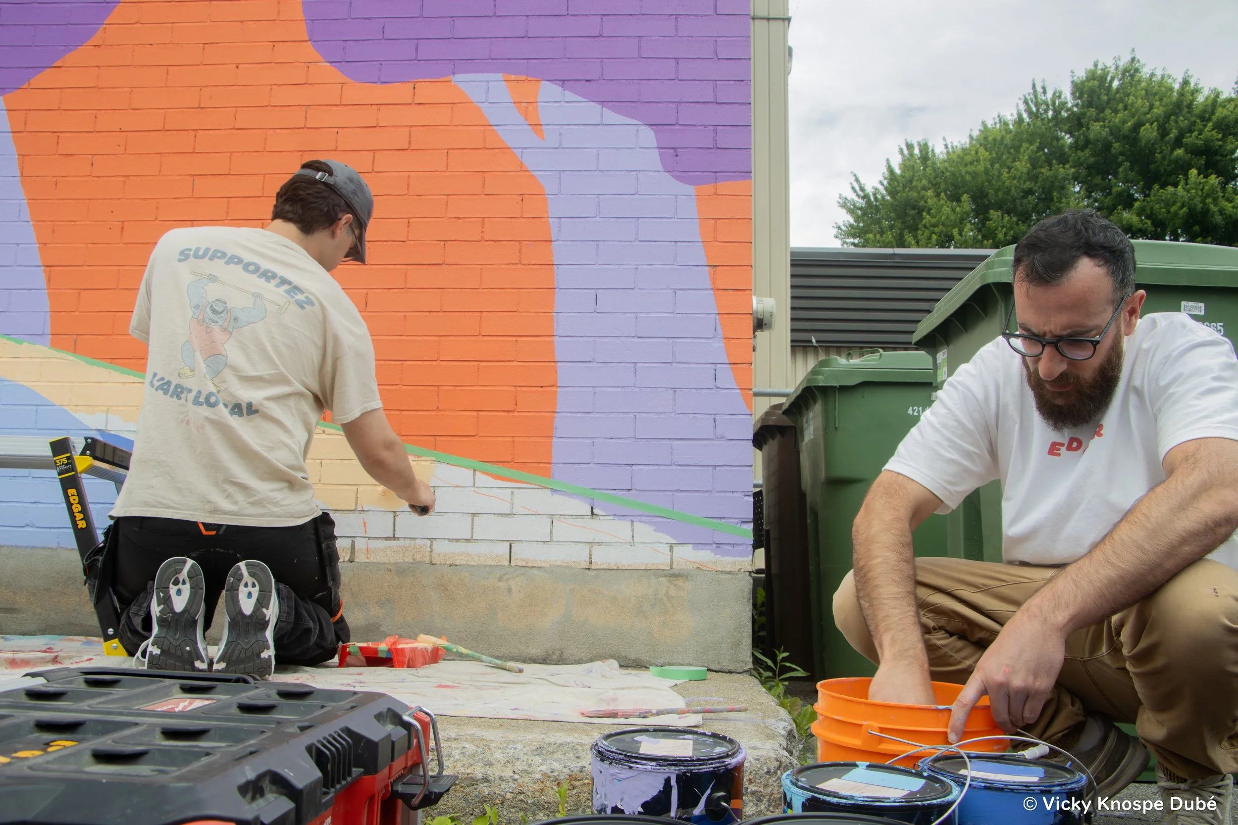 Deux hommes peignant une fresque murale sur un mur en briques, avec un seau de peinture et des outils à proximité.