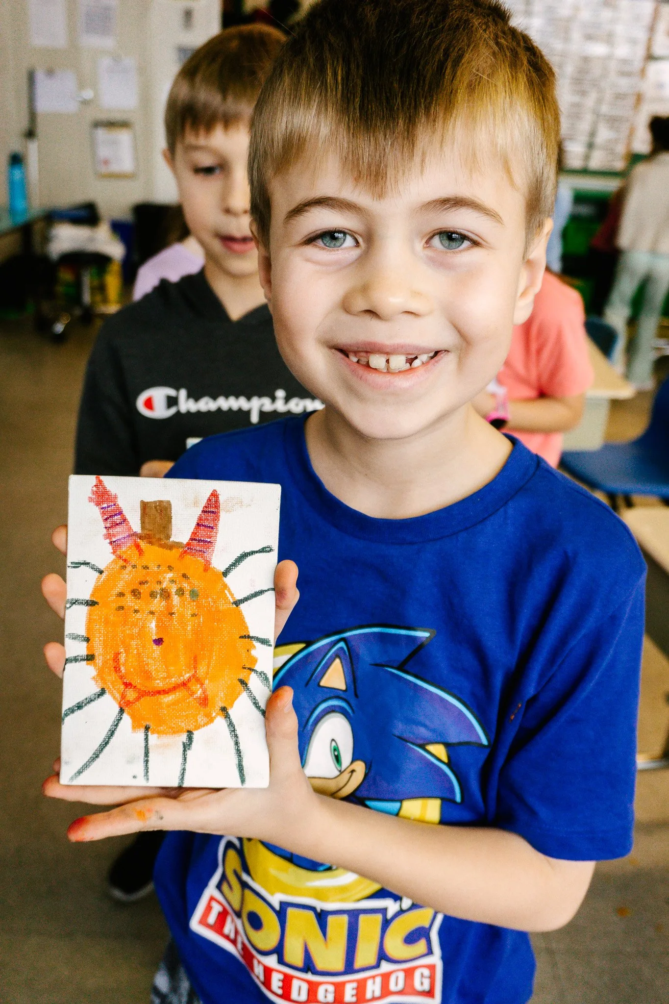 Enfant tenant un dessin d'un visage orange souriant, avec des oreilles rouges et un encadrement noir de cheveux. En arrière-plan, un autre enfant et une salle de classe. Atelier fait par edgar artiste peintre pour l'école primaire jean 23