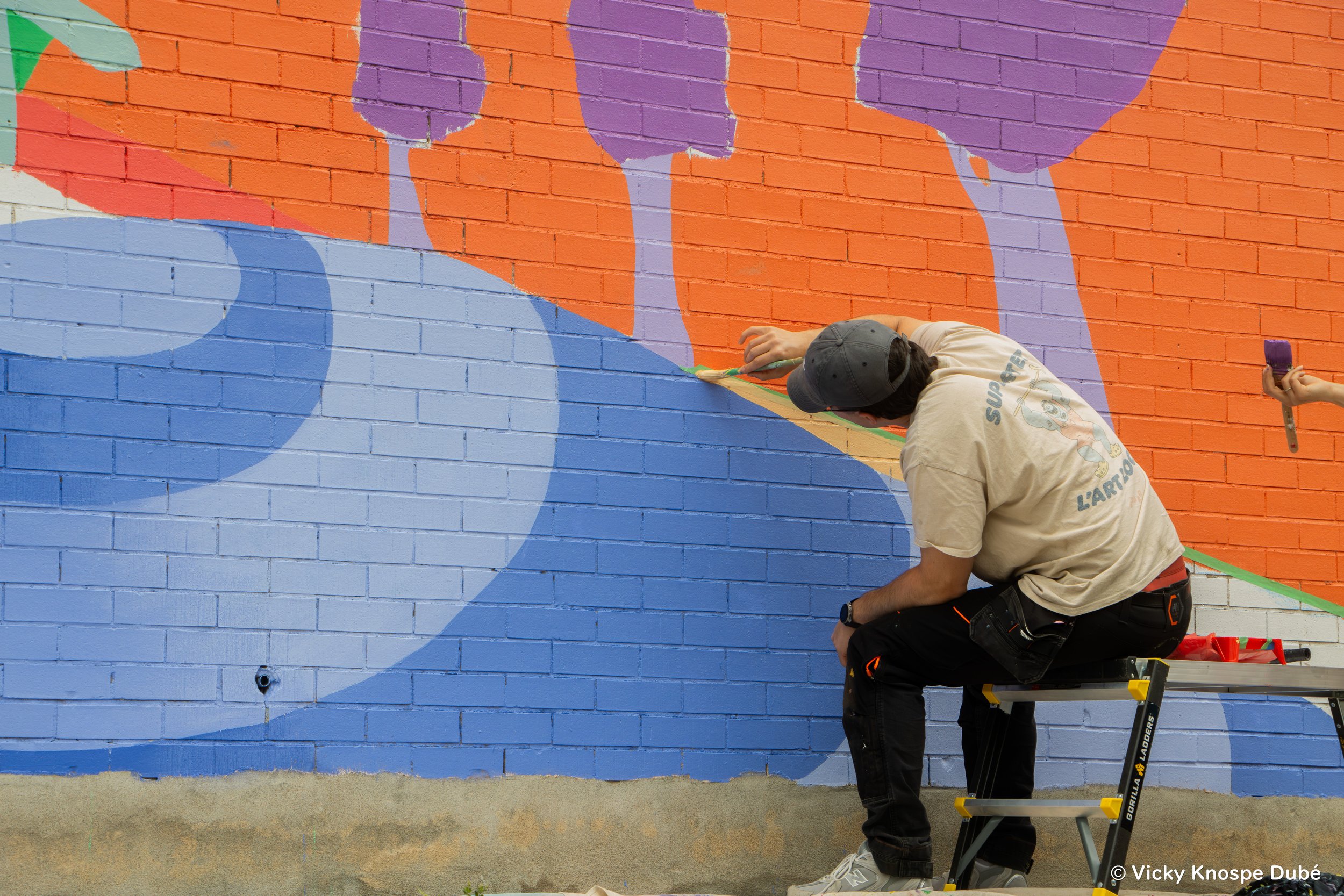Un artiste qui peint une fresque murale colorée sur un mur en briques orange et bleue avec un escabeau. La fresque comporte des formes abstraites dans des tons violets et pastels.