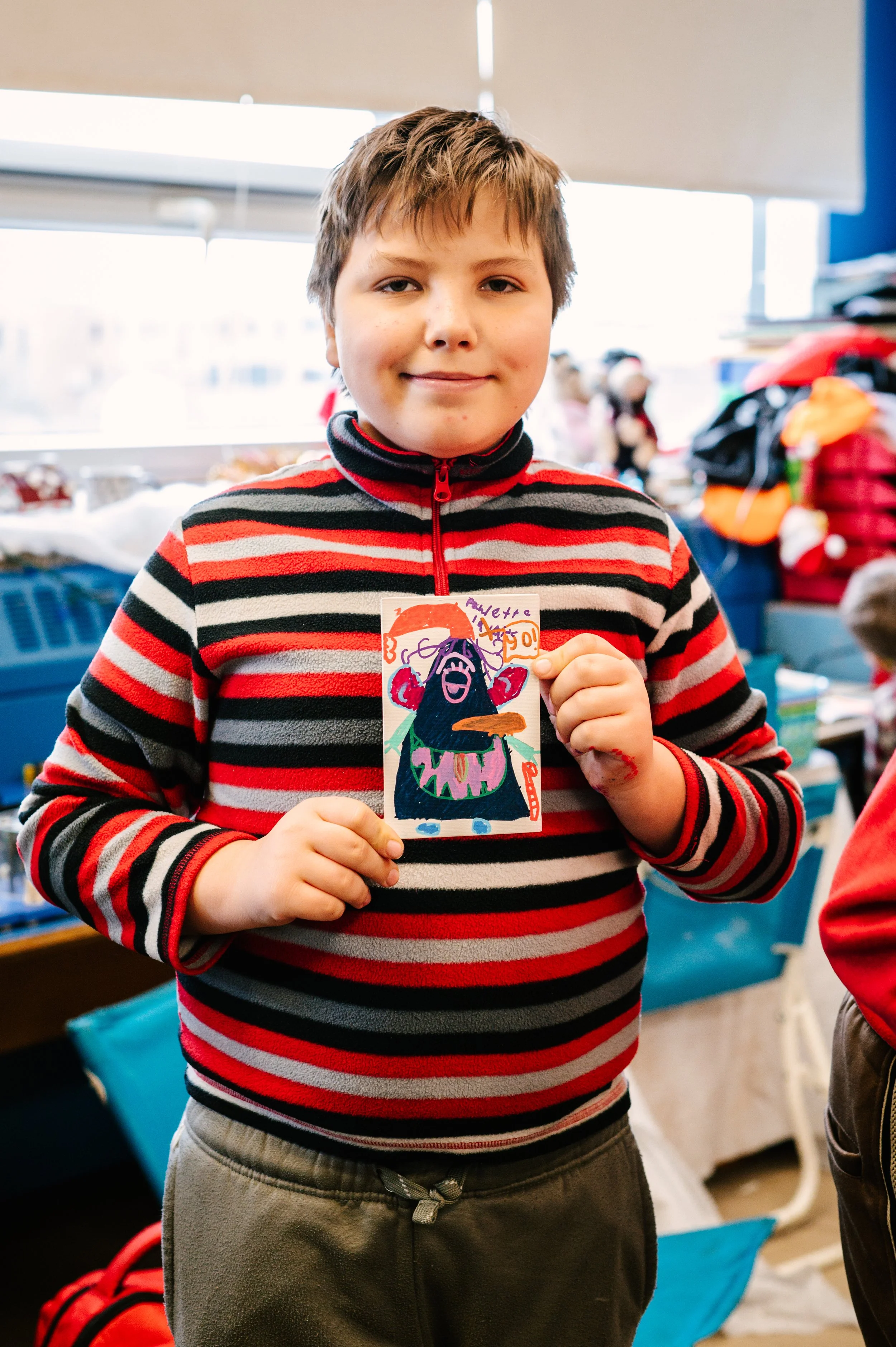 Un enfant en pull rayé rouge et noir tient une carte dessinée à la main dans une salle de classe. Atelier fait par edgar artiste peintre pour l'école primaire jean 23