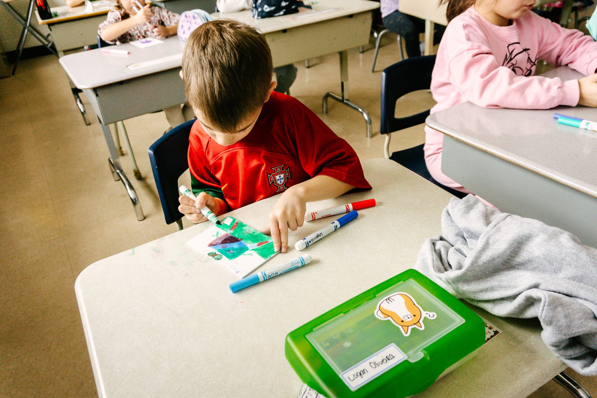 Enfant dessinant à l'école, assis à un bureau avec des feutres colorés et une boîte verte sur la table. Atelier fait par edgar artiste peintre pour l'école primaire jean 23