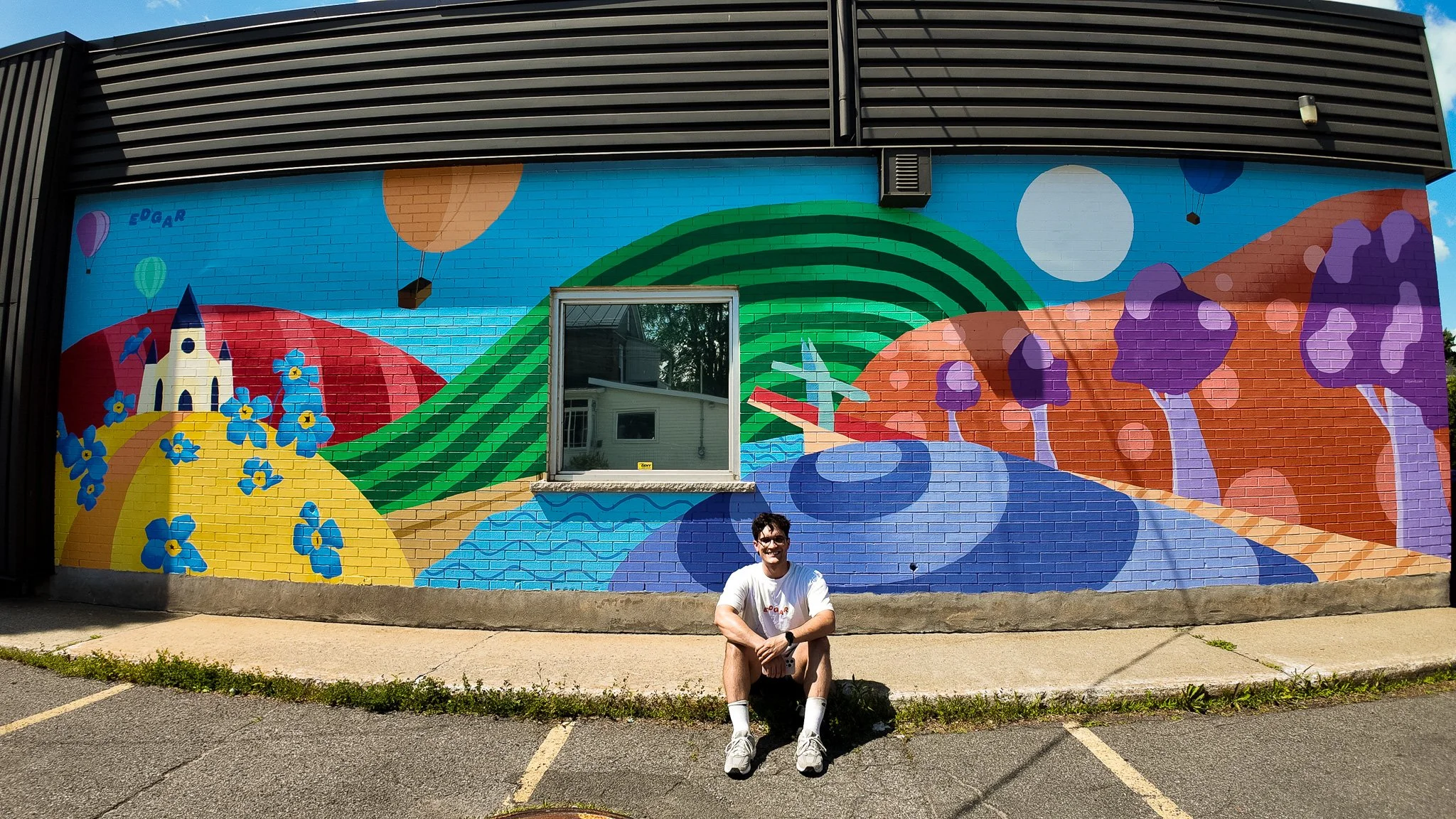 Mural coloré représentant un paysage avec une maison, des collines, des arbres violets et un ciel avec des ballons, et un homme assis devant.