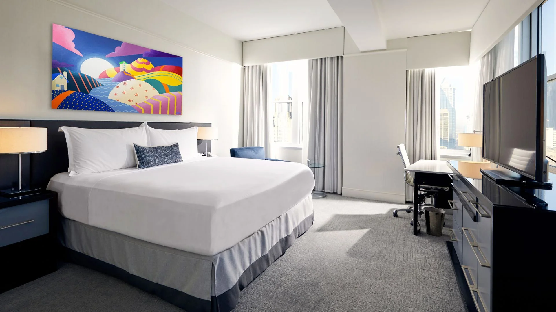 Chambre d'hôtel moderne et lumineuse avec un grand lit double, deux lampes de chevet, un tableau coloré sur le mur, un bureau avec une chaise, une grande télévision à écran plat, et une vue sur des immeubles par la fenêtre.