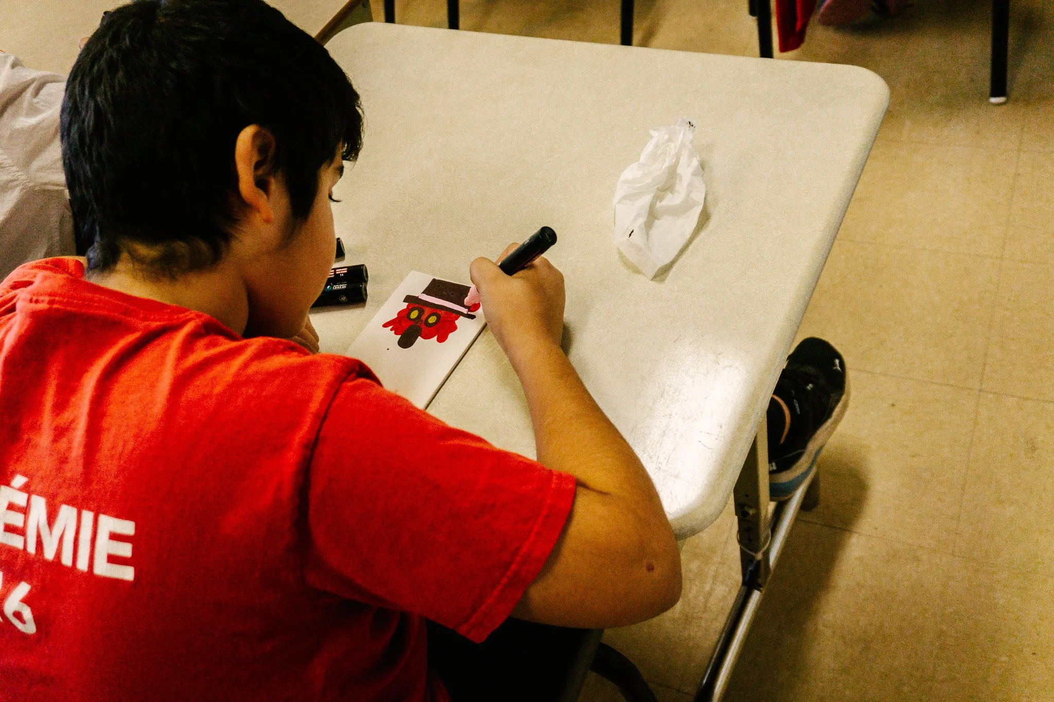Enfant en train de dessiner sur une feuille avec un stylo, portant un t-shirt rouge, assis à une table dans une salle de classe. Atelier fait par edgar artiste peintre pour l'école primaire jean 23