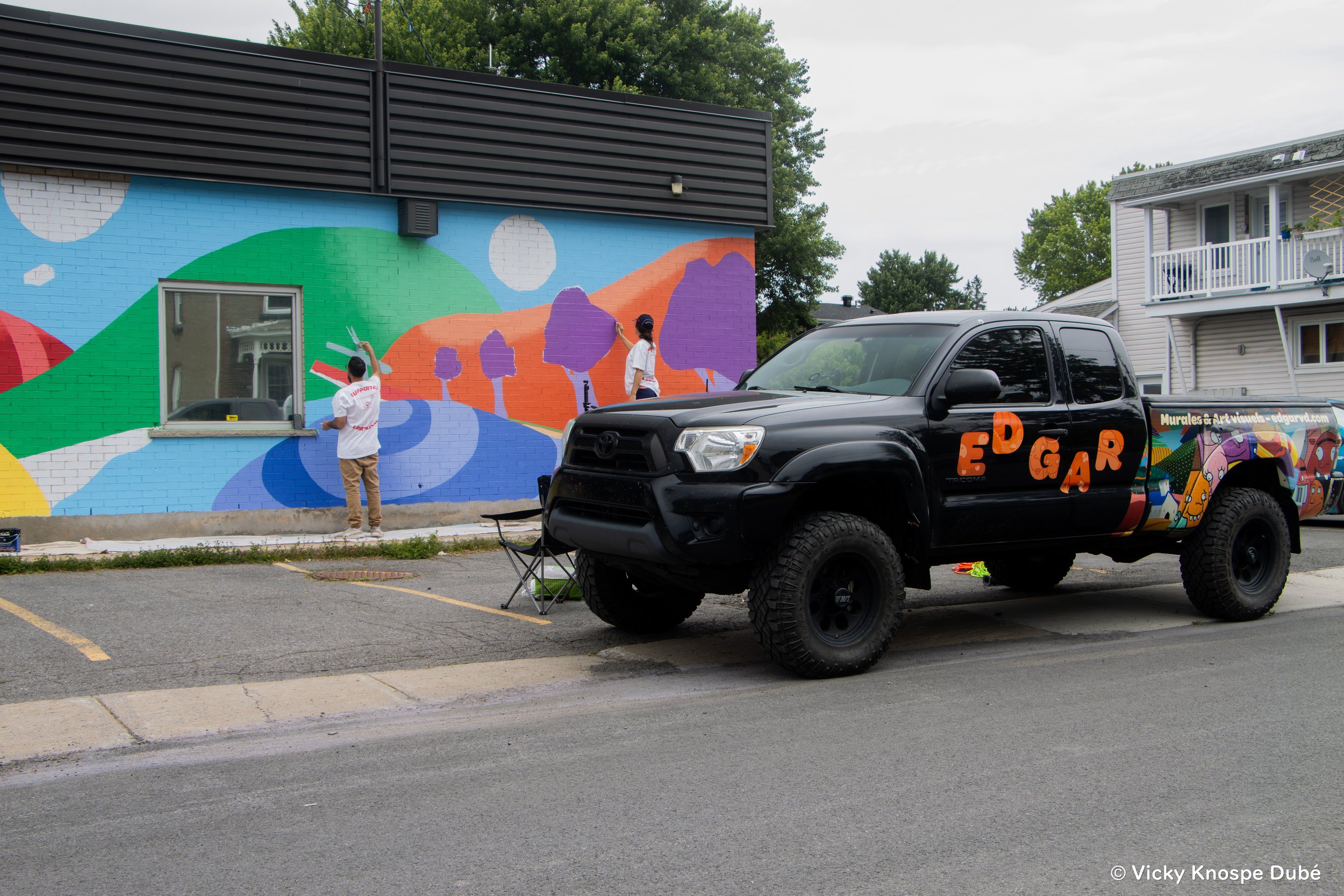 Deux artistes peignent une fresque murale colorée sur un bâtiment, avec un camion noir à côté portant le nom 'EDGAR' en lettres orange.
