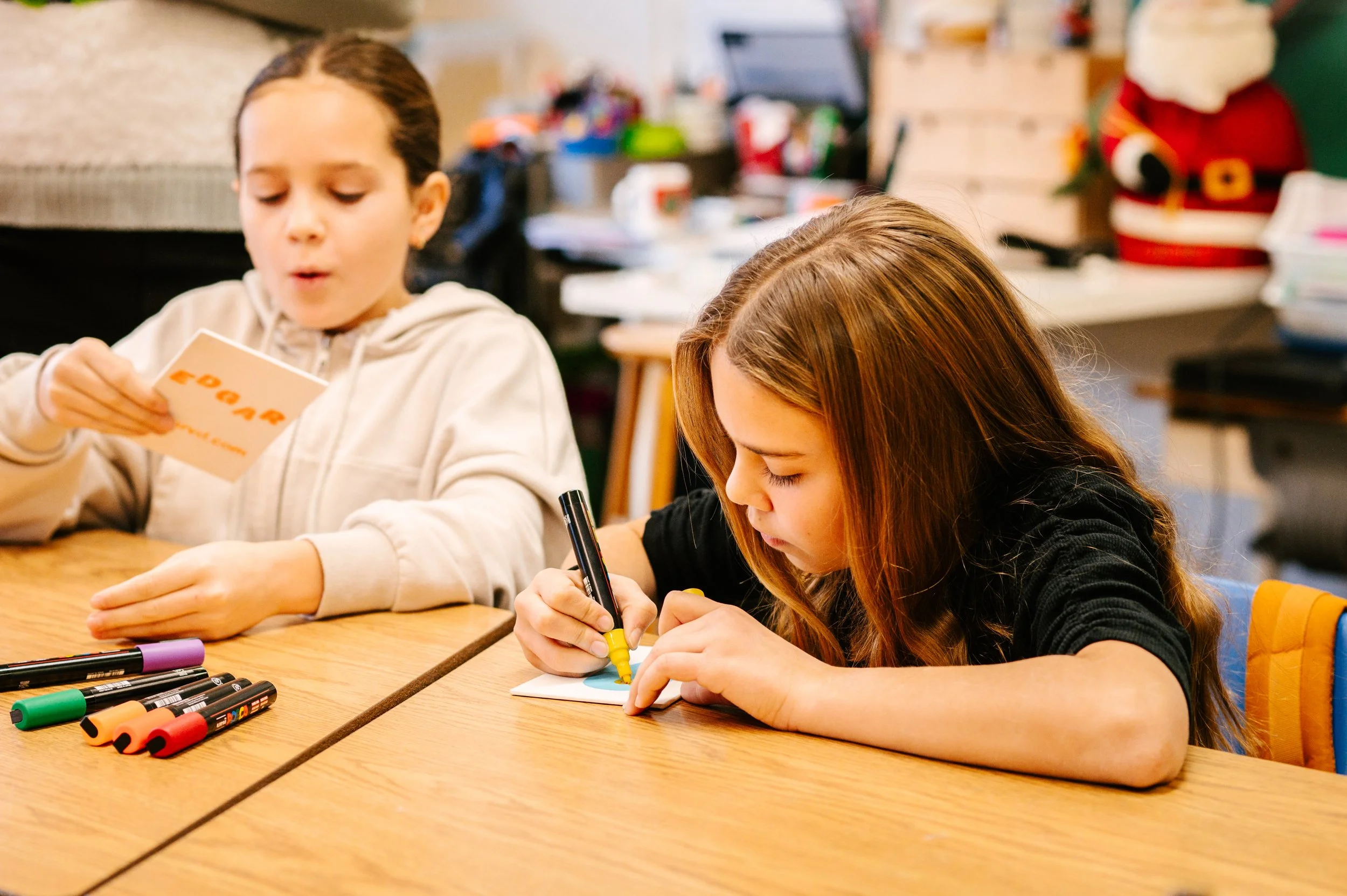 Deux enfants dans une salle de classe, l'un écrivant et l'autre regardant une feuille de papier, avec des marqueurs colorés sur la table. Atelier fait par edgar artiste peintre pour l'école primaire jean 23