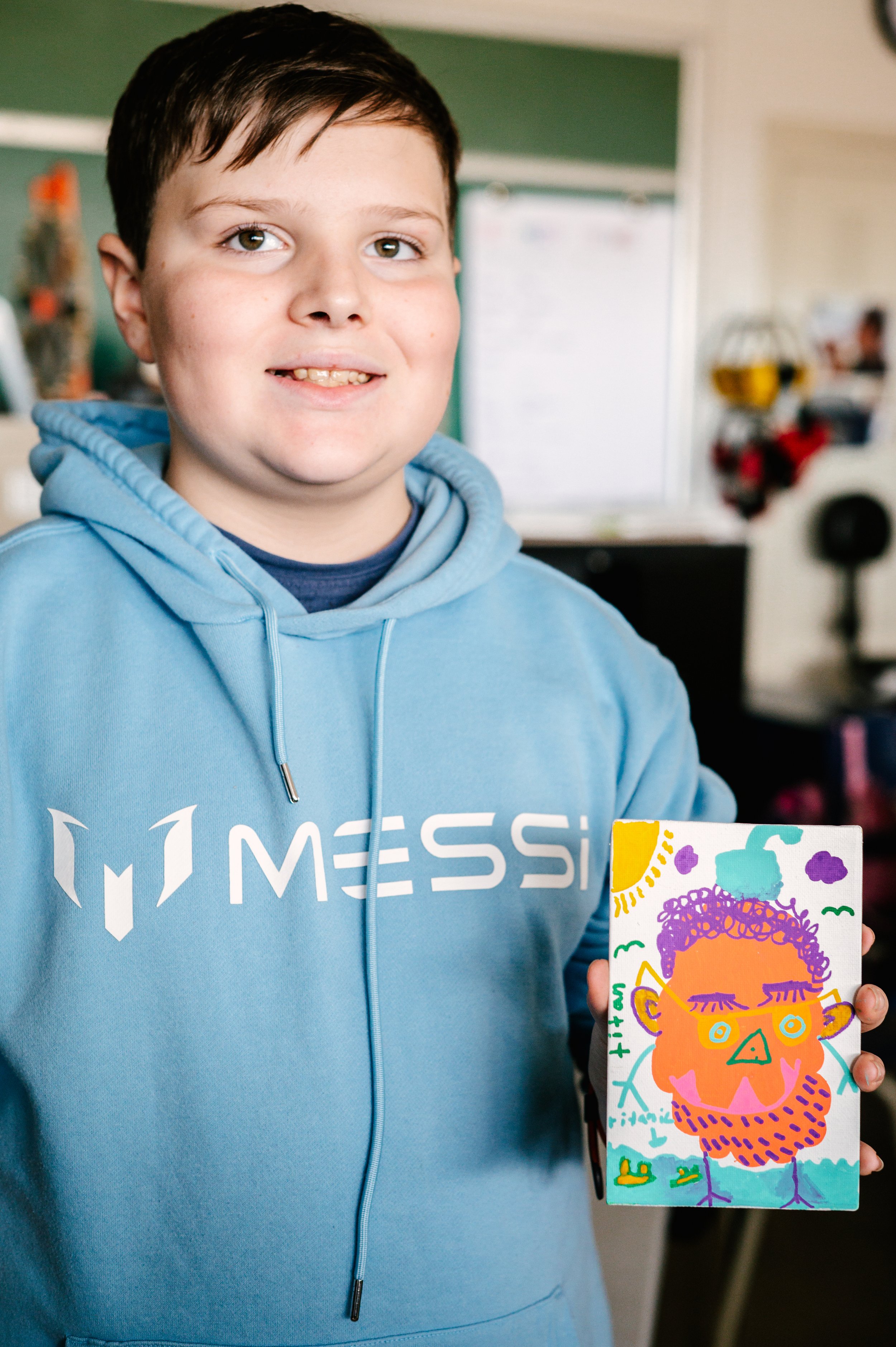 Un enfant portant un sweat à capuche bleu avec l'inscription "Messi", tenant un dessin coloré d'un visage stylisé et abstrait. Atelier fait par edgar artiste peintre pour l'école primaire jean 23