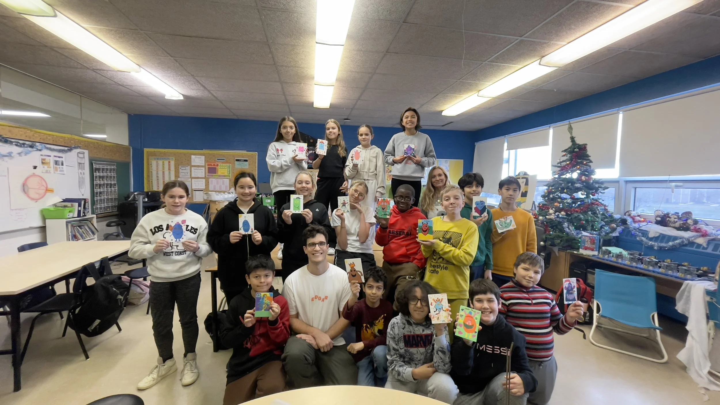 Groupe d'enfants et d'adultes dans une salle de classe, tenant des cartes faites à la main devant un sapin de Noël décoré. Atelier fait par edgar artiste peintre pour l'école primaire jean 23