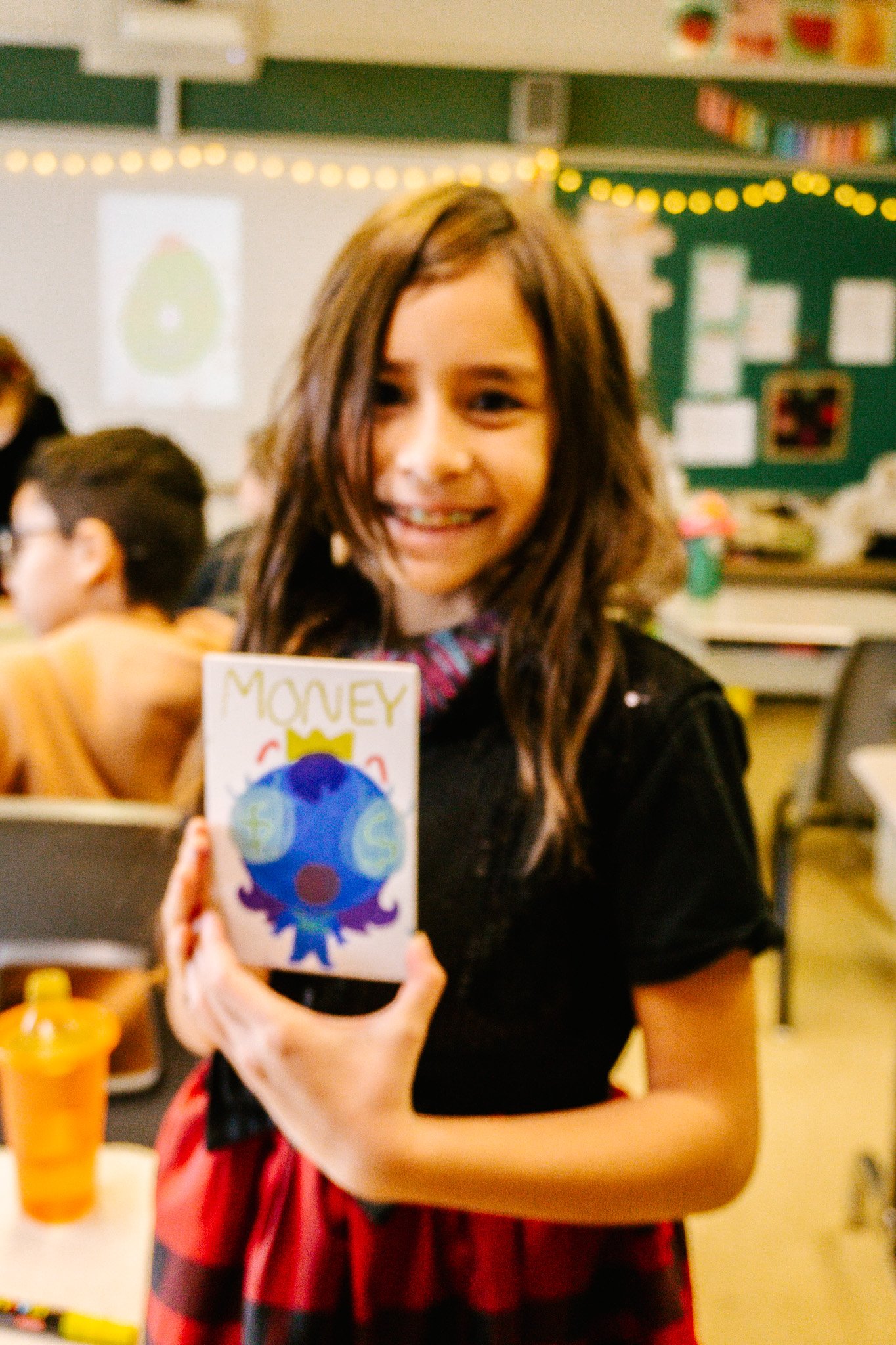 Fille souriante tenant un dessin avec "MONEY" écrit dessus dans une salle de classe. Atelier fait par edgar artiste peintre pour l'école primaire jean 23