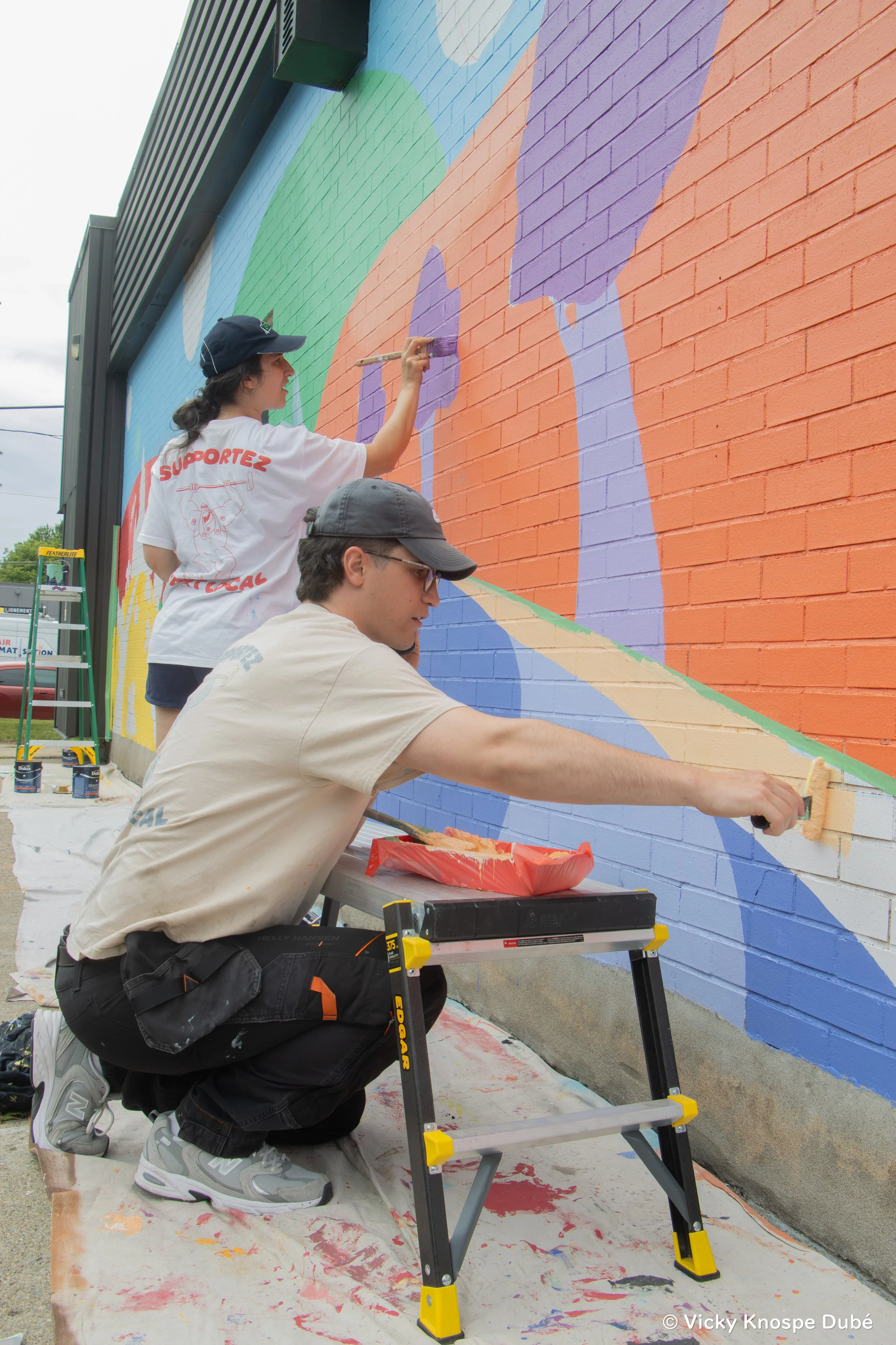 Deux jeunes artistes peignent une fresque murale colorée sur un mur en briques, à l'extérieur.