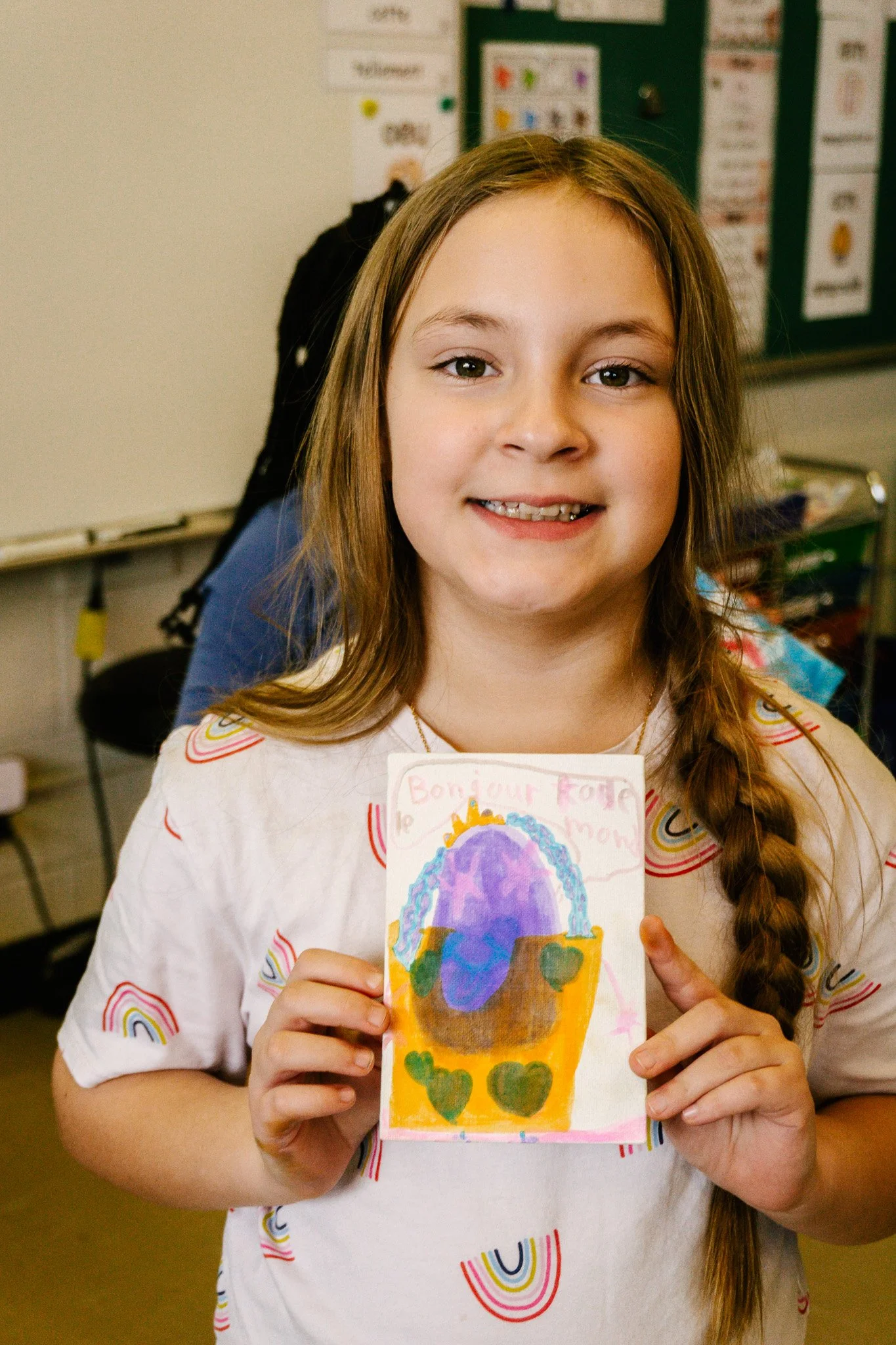 Fille souriante tenant un dessin coloré avec des cœurs et des écritures, dans une salle de classe. Atelier fait par edgar artiste peintre pour l'école primaire jean 23
