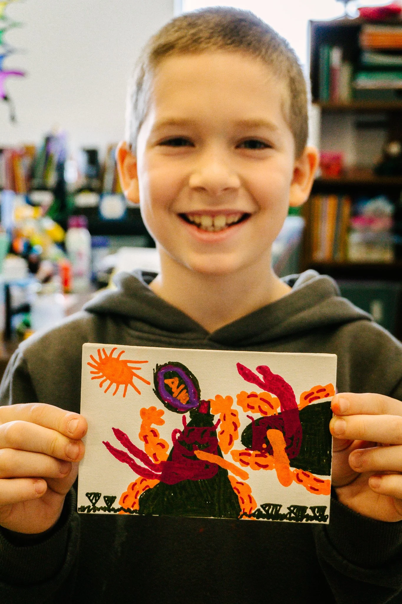 Un enfant souriant tenant un dessin coloré représentant un paysage avec des formes abstraites et un soleil orange. Atelier fait par edgar artiste peintre pour l'école primaire jean 23