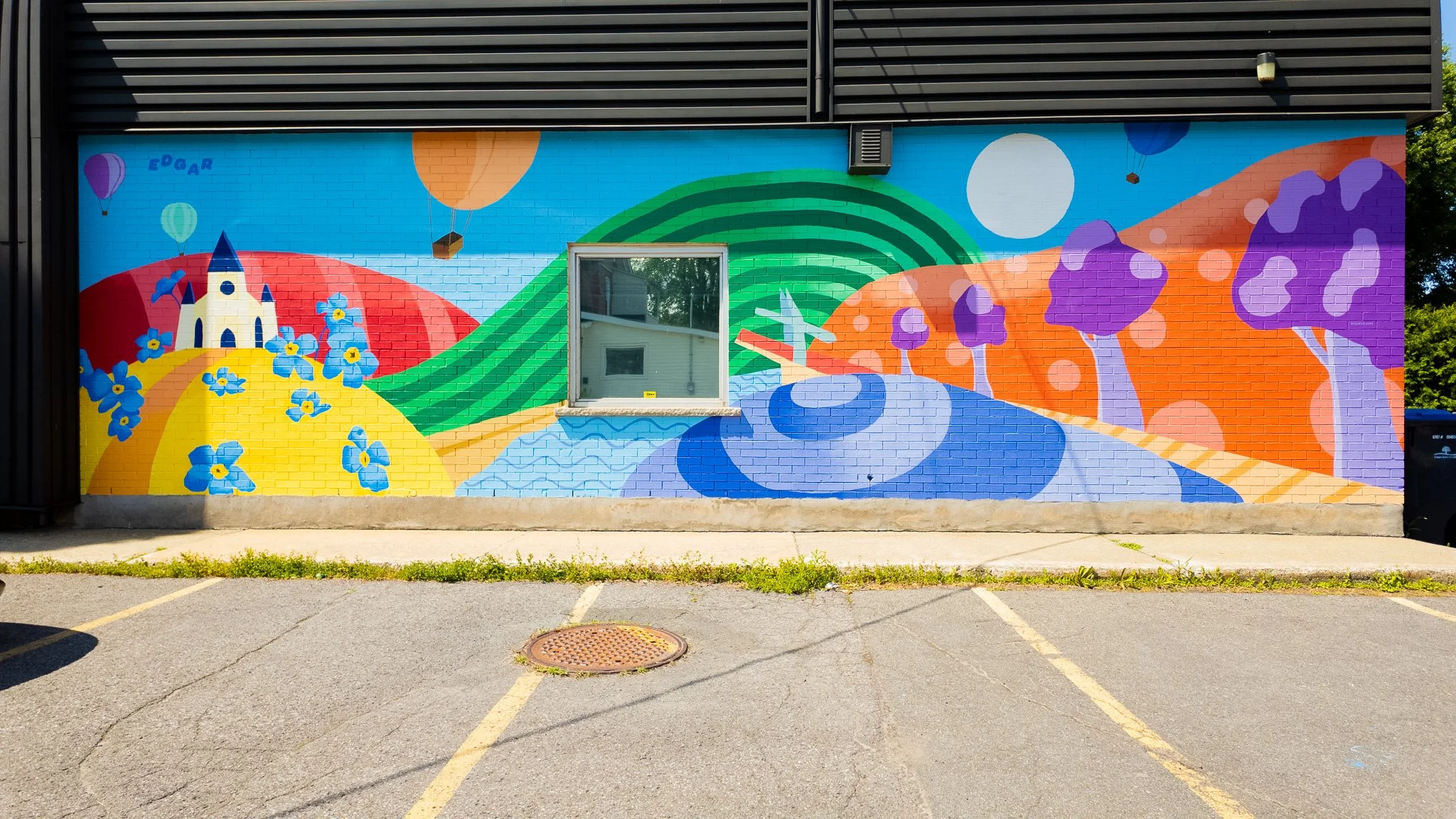 Fresque murale colorée représentant un paysage fantastique avec des collines, un château, des arbres violets, un soleil, des ballons, et un moulin à vent, avec une fenêtre au centre.