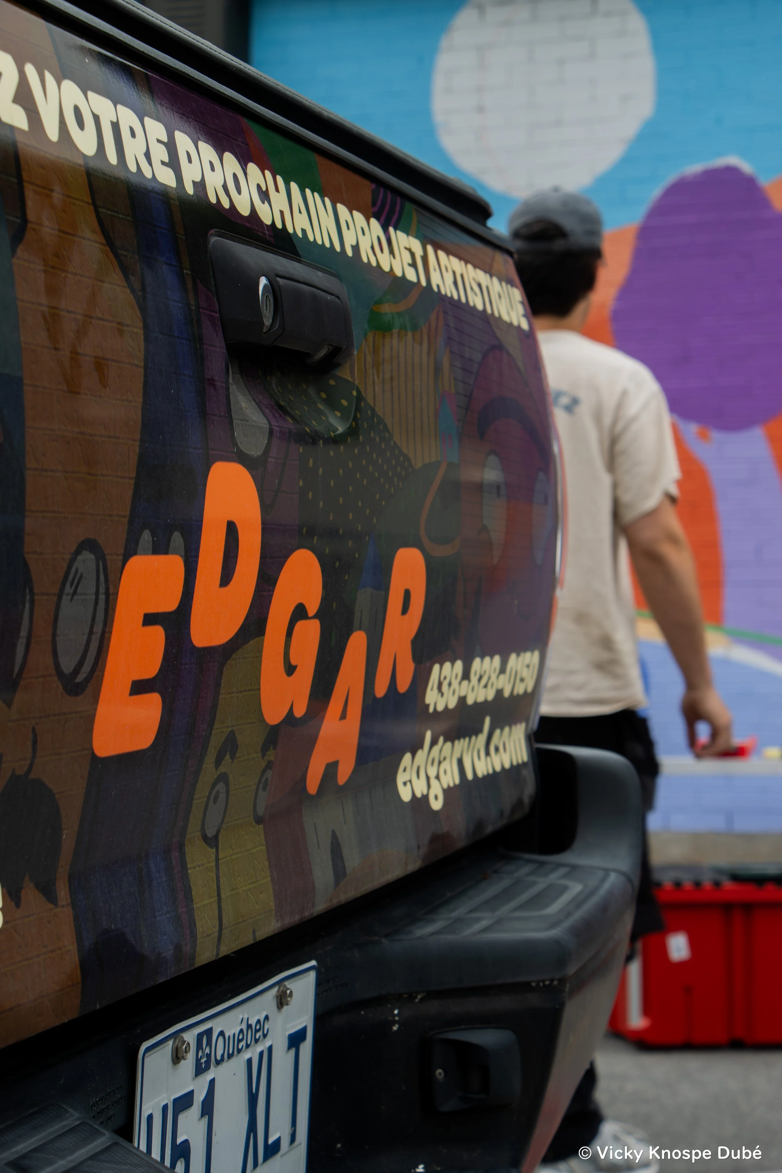 Une camionnette avec un panneau promotionnel orange et violet pour un projet artistique au Québec, avec un homme en arrière-plan devant un mur peint en bleu et violet