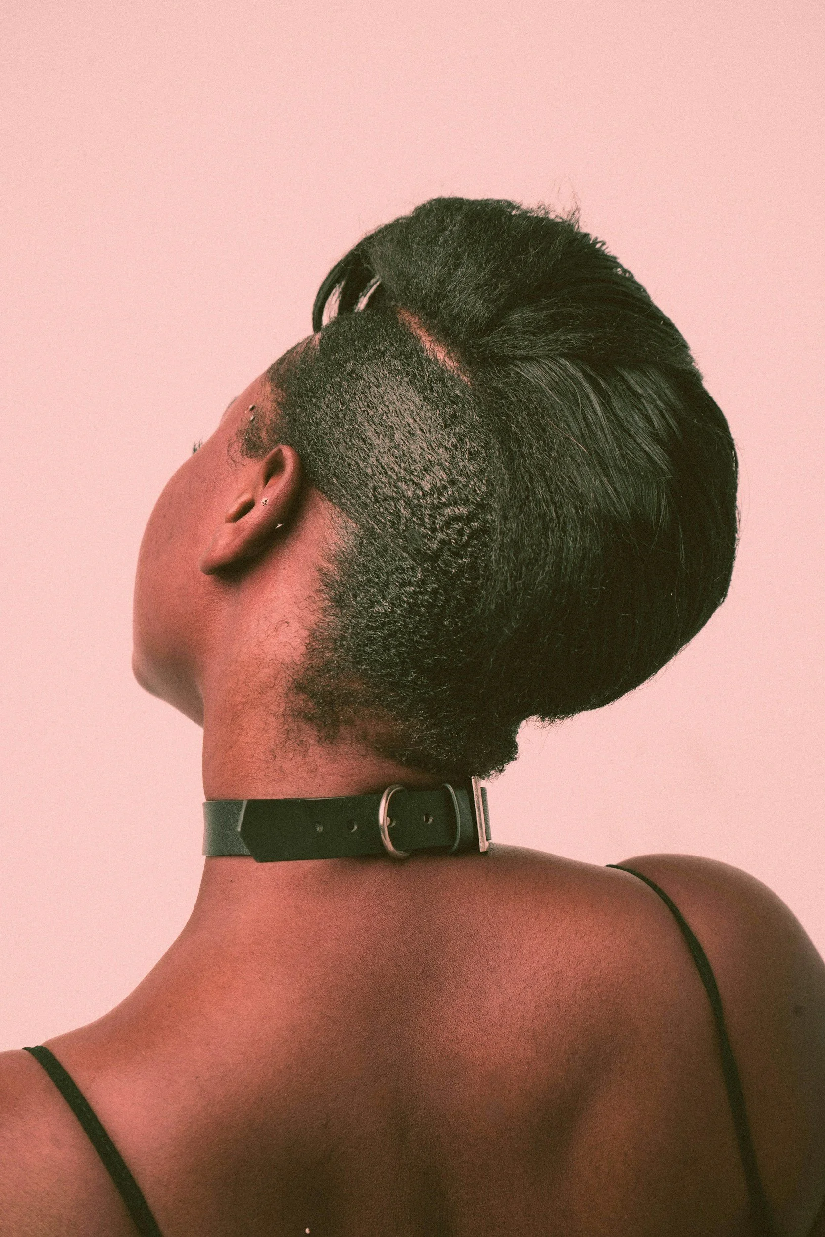 a Black woman with short hair is wearing a black leather submissive collar bucked at the back of her neck