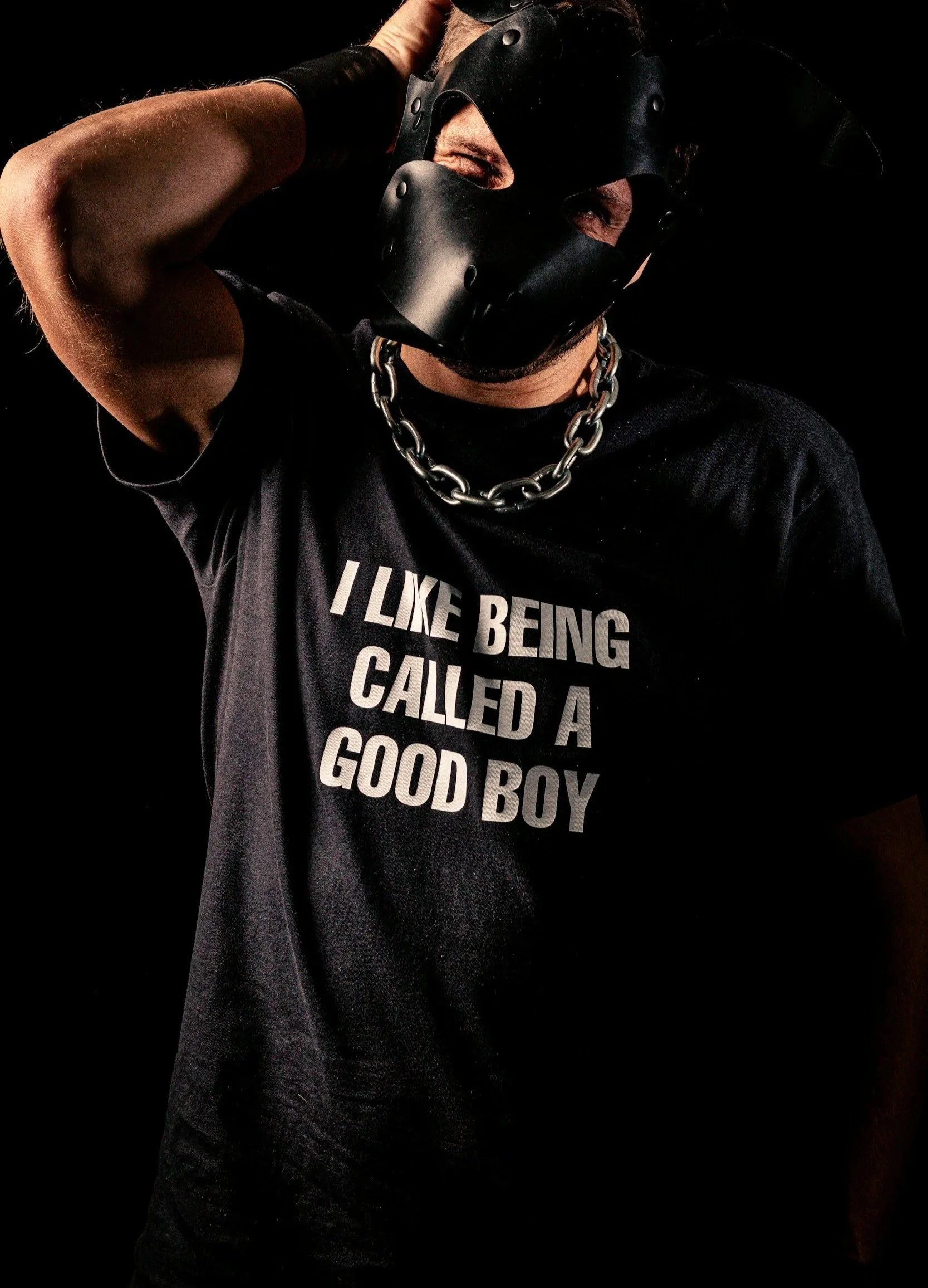 a male submissive wearing a pup play mask and a chain collar with a t-shirt that says "I like being called a good boy"
